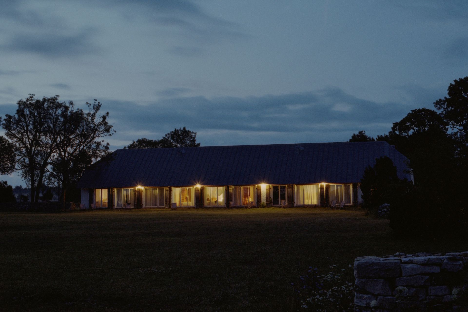 Long, lit building at dusk, windows lit, against a dark sky. A stone wall is in the foreground.