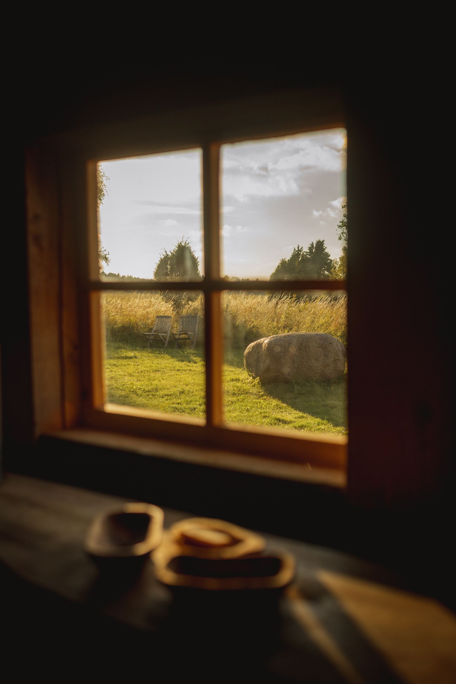 View through a window of a grassy field with trees and a setting sun. Small plates on a wooden ledge.