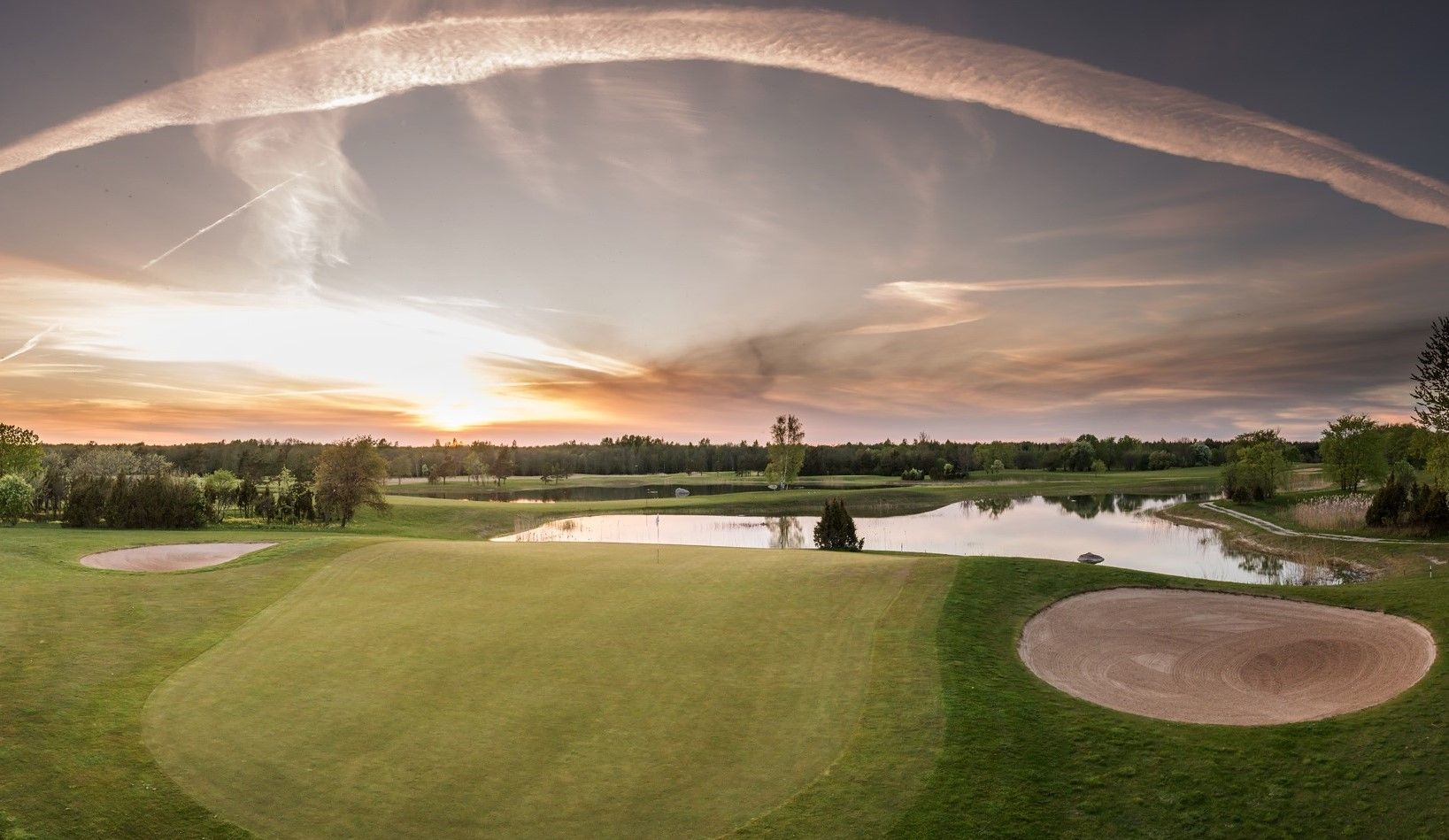 Golf course at sunset with green grass, lake, sand traps, and dramatic sky.
