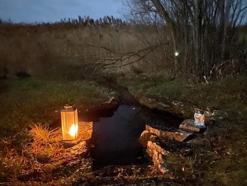 A glowing lantern and candle flank a dark pool, set in a grassy field with trees at dusk.