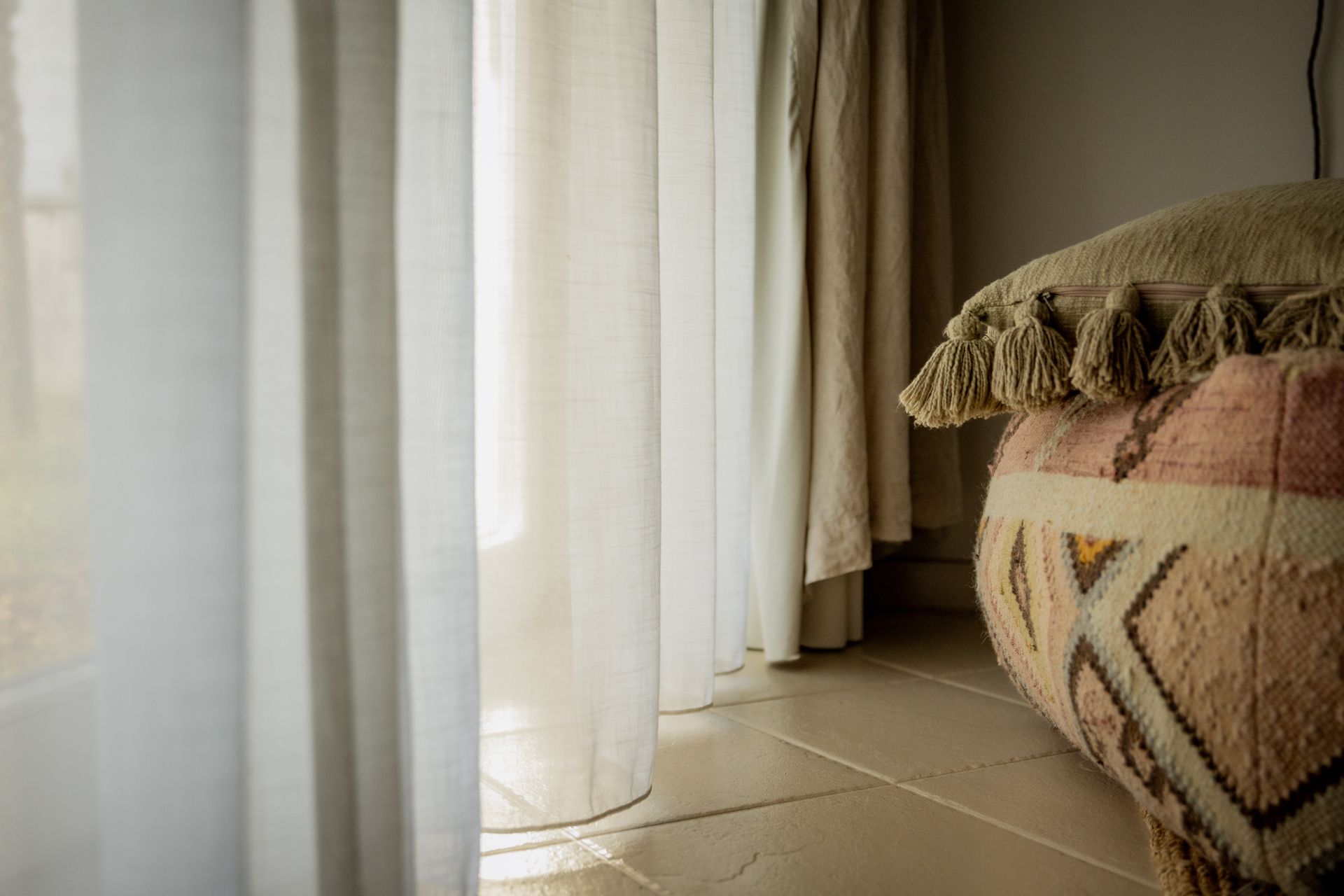 Sheer white curtains next to a decorative patterned pouf in a room with tiled floors.