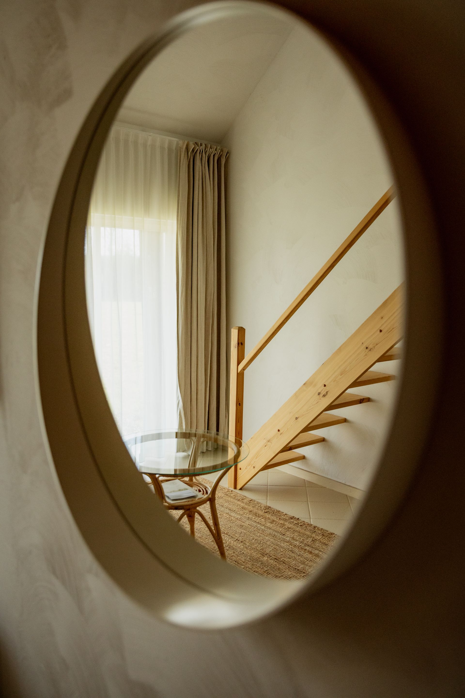 Round mirror reflecting a room: stairs, table with glass top, curtains, and neutral-colored walls.