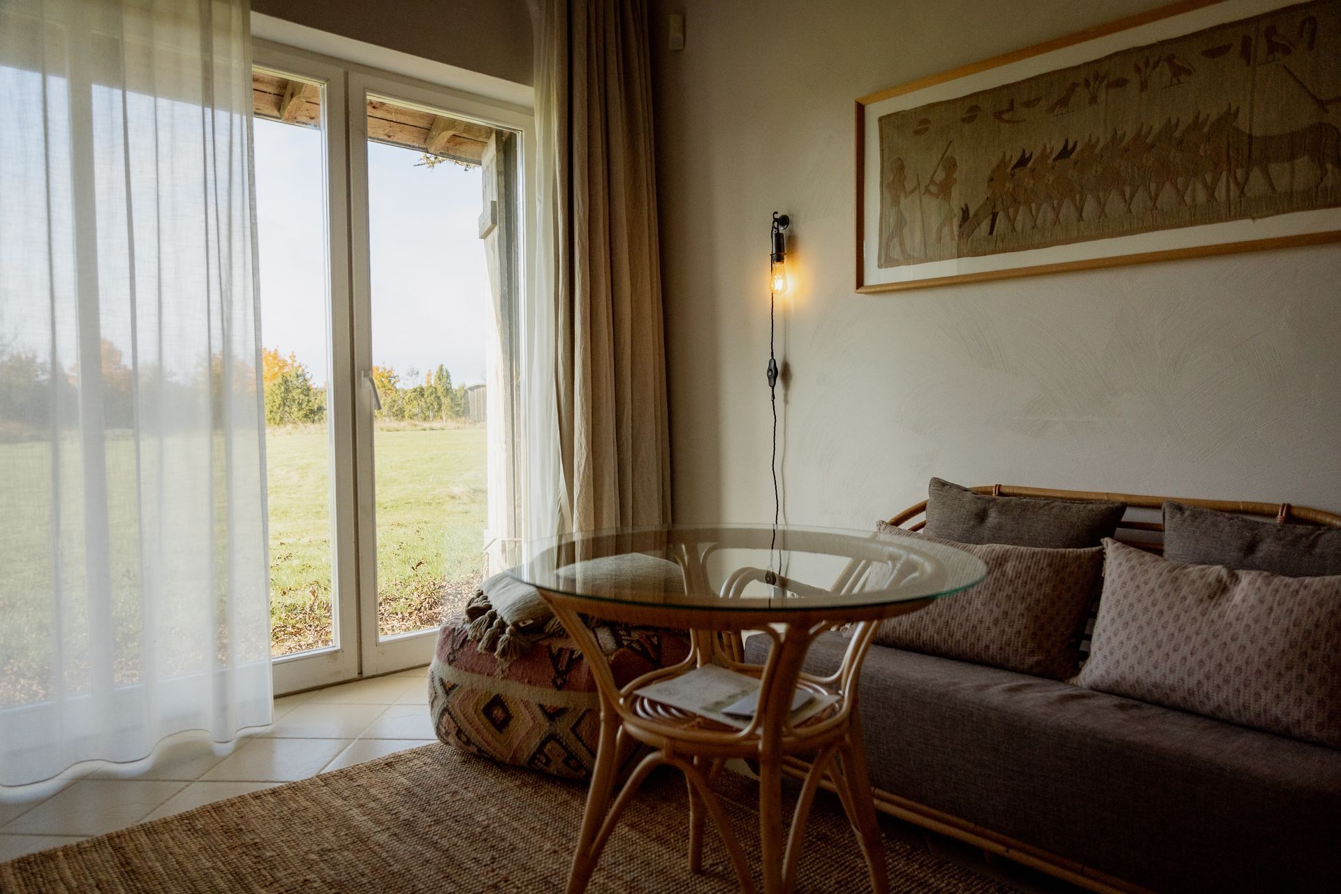 Interior of a room with a view: Glass table, wicker chair, sofa with pillows, large window with sheer curtains.