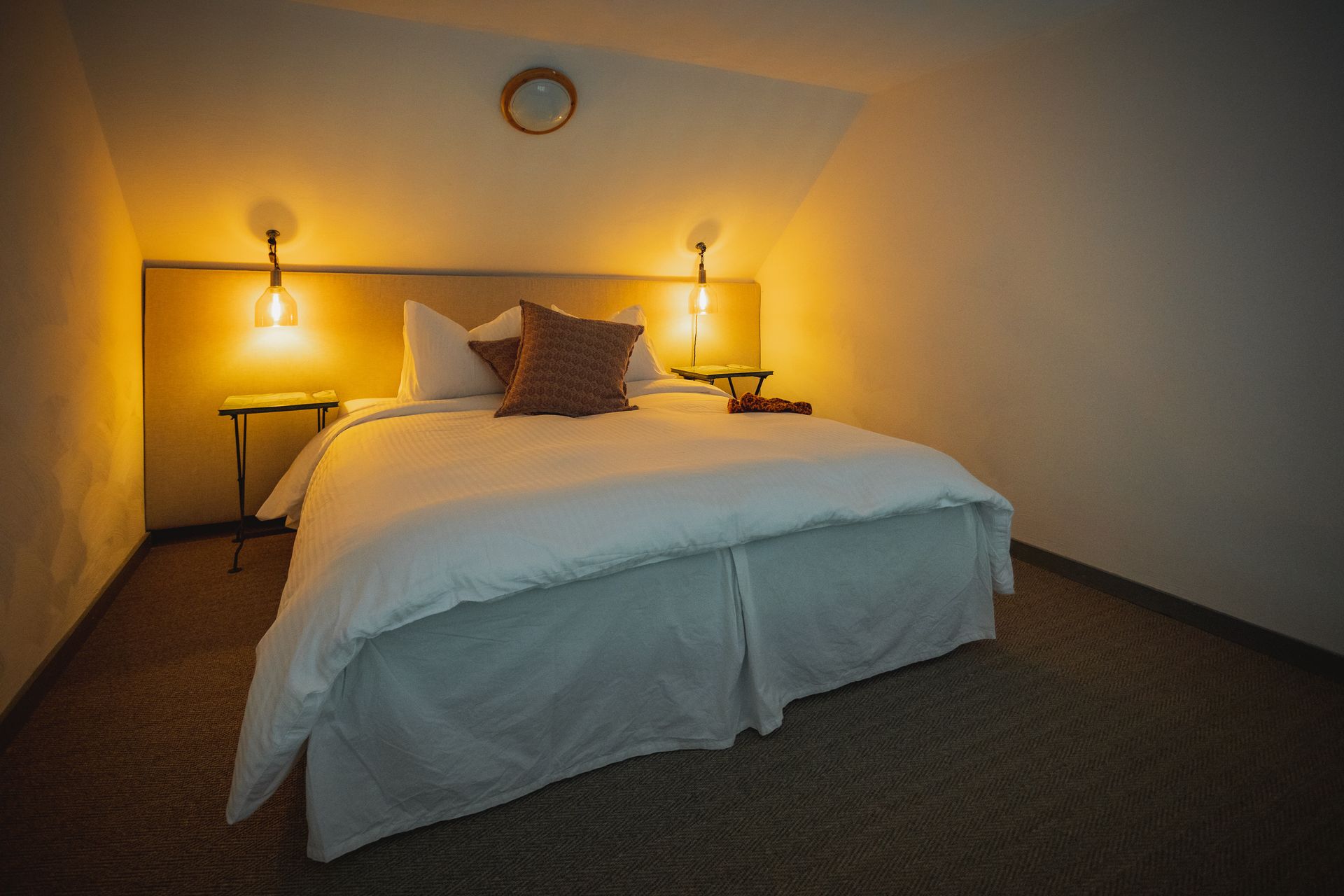 Bedroom with bed, side tables, lamps, and angled ceiling. Warm lighting.