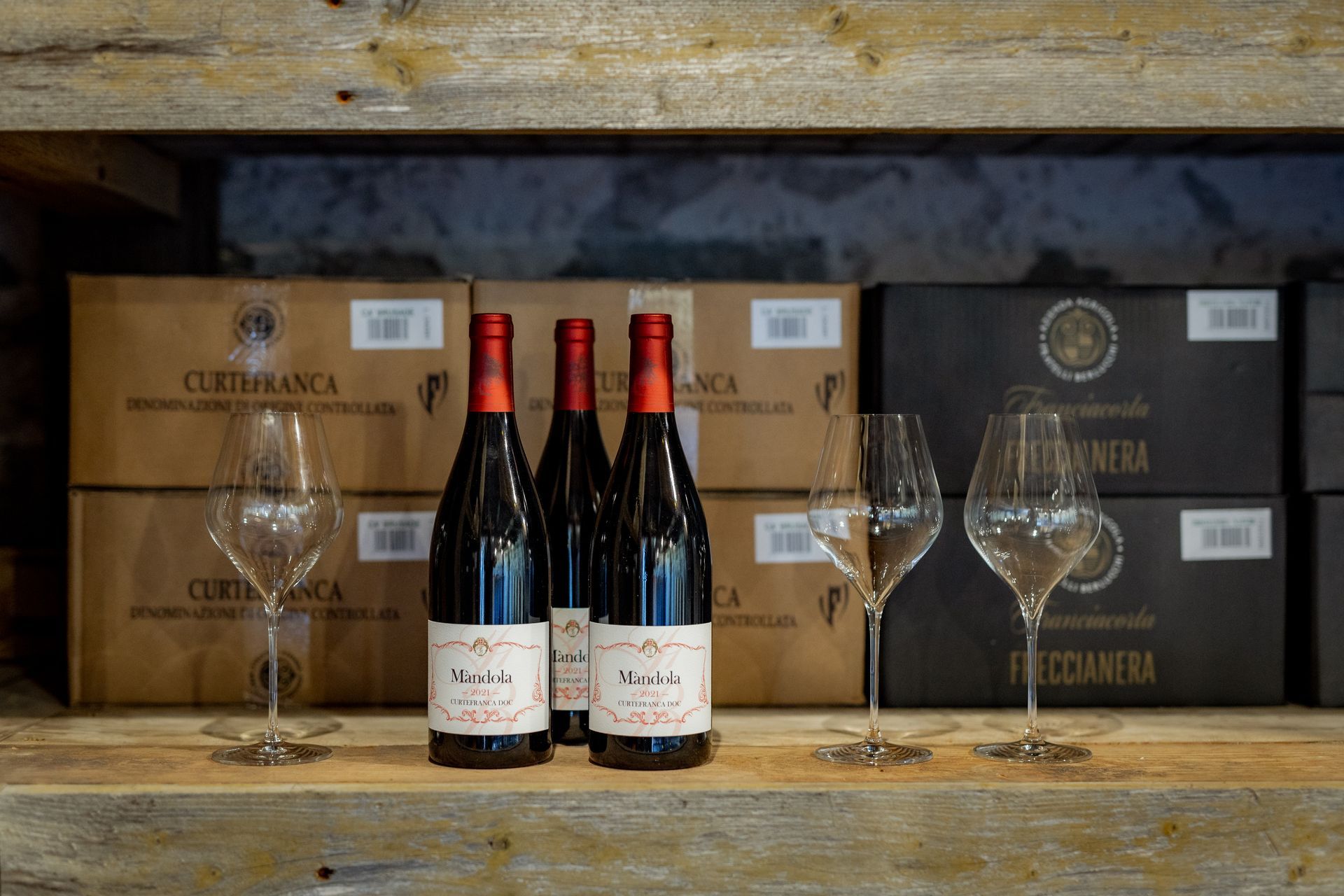 Three bottles of red wine and two wine glasses on a wooden shelf in front of boxes.