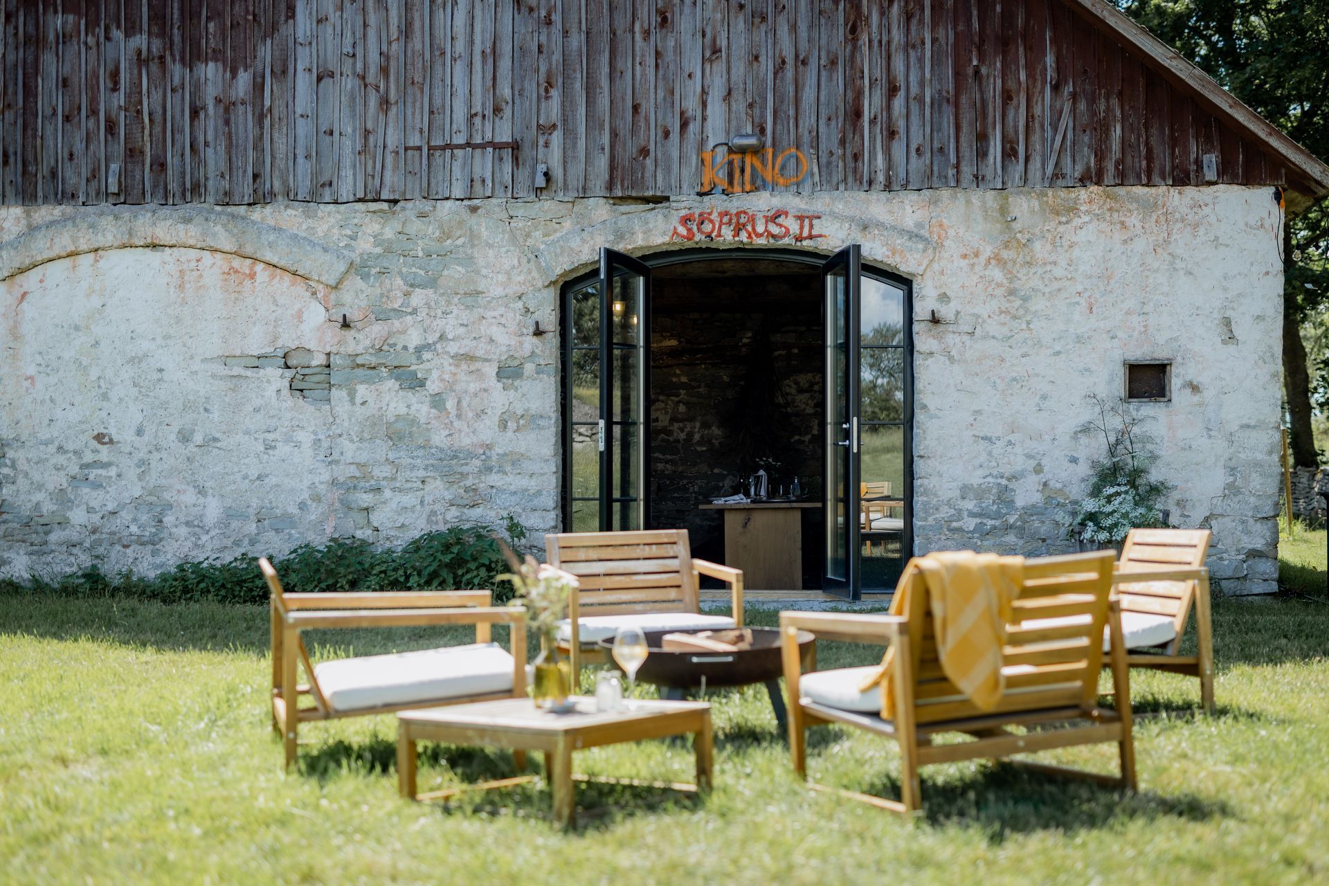 Outdoor seating area with wooden furniture, in front of a weathered stone building with open doors.