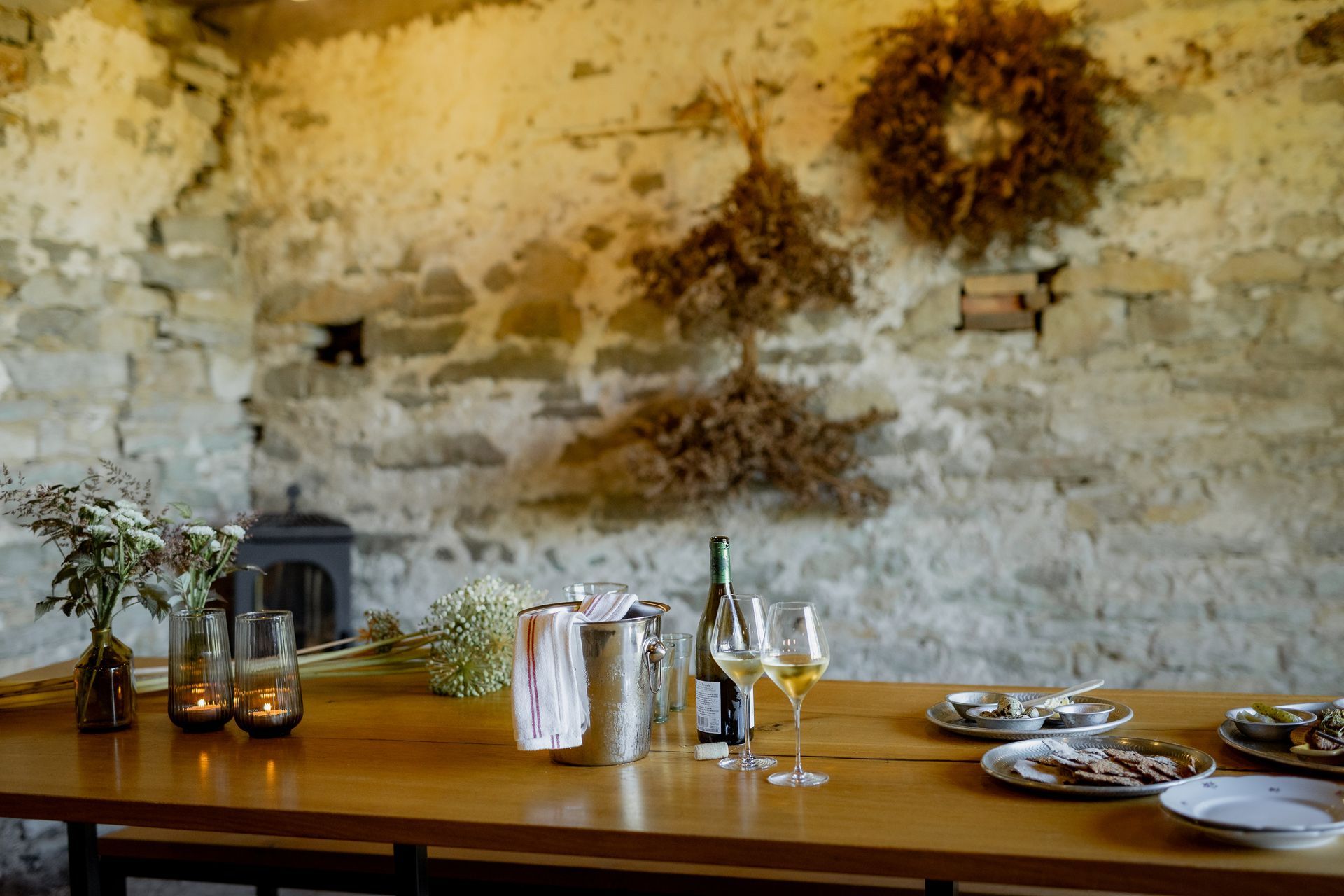 Wooden table set for a meal in a stone-walled room, with wine, plates, flowers, and decorations.