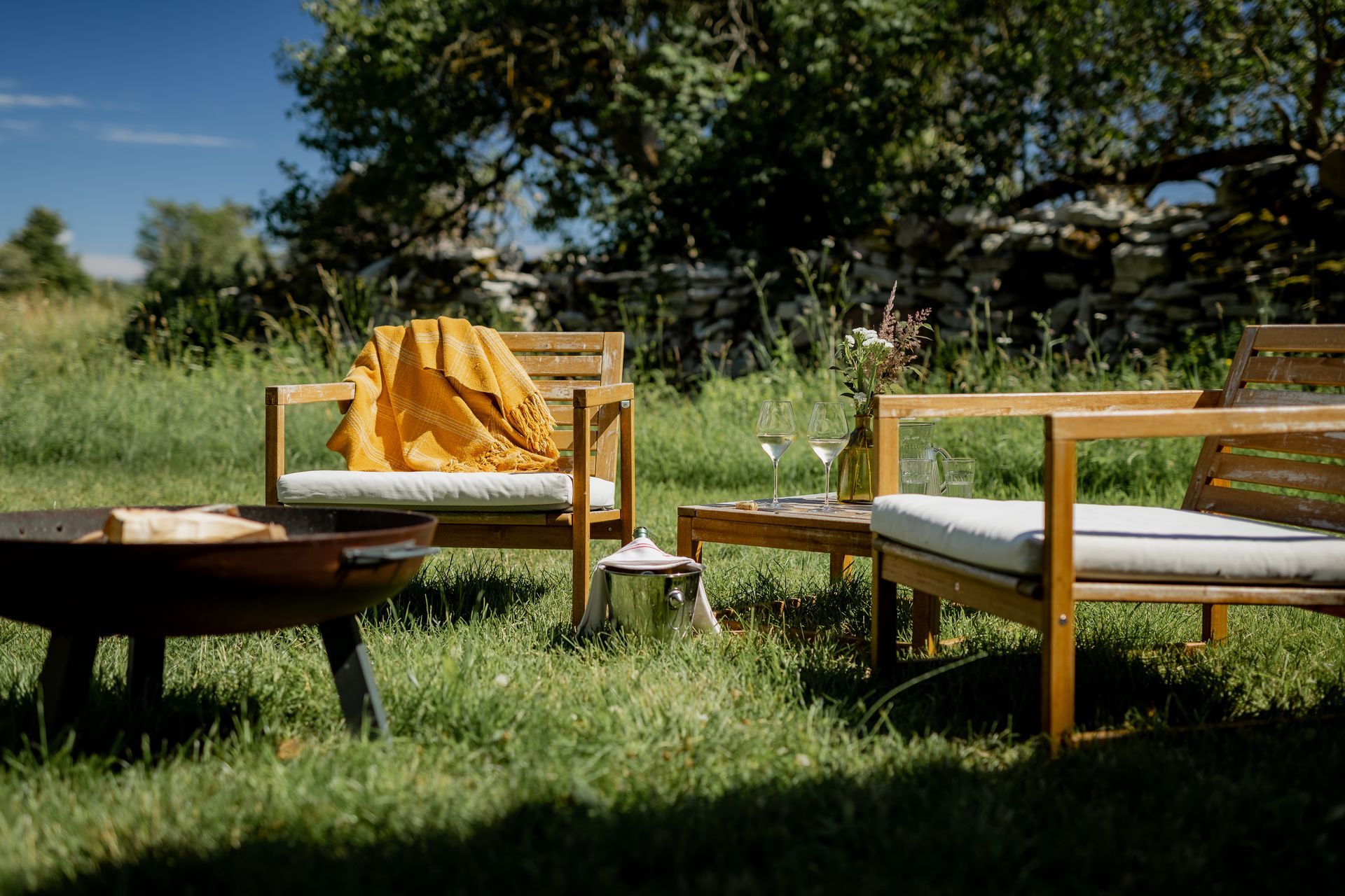 Outdoor seating area: wooden chairs with cushions, small table with drinks, blanket, fire pit, green grass, sunny.