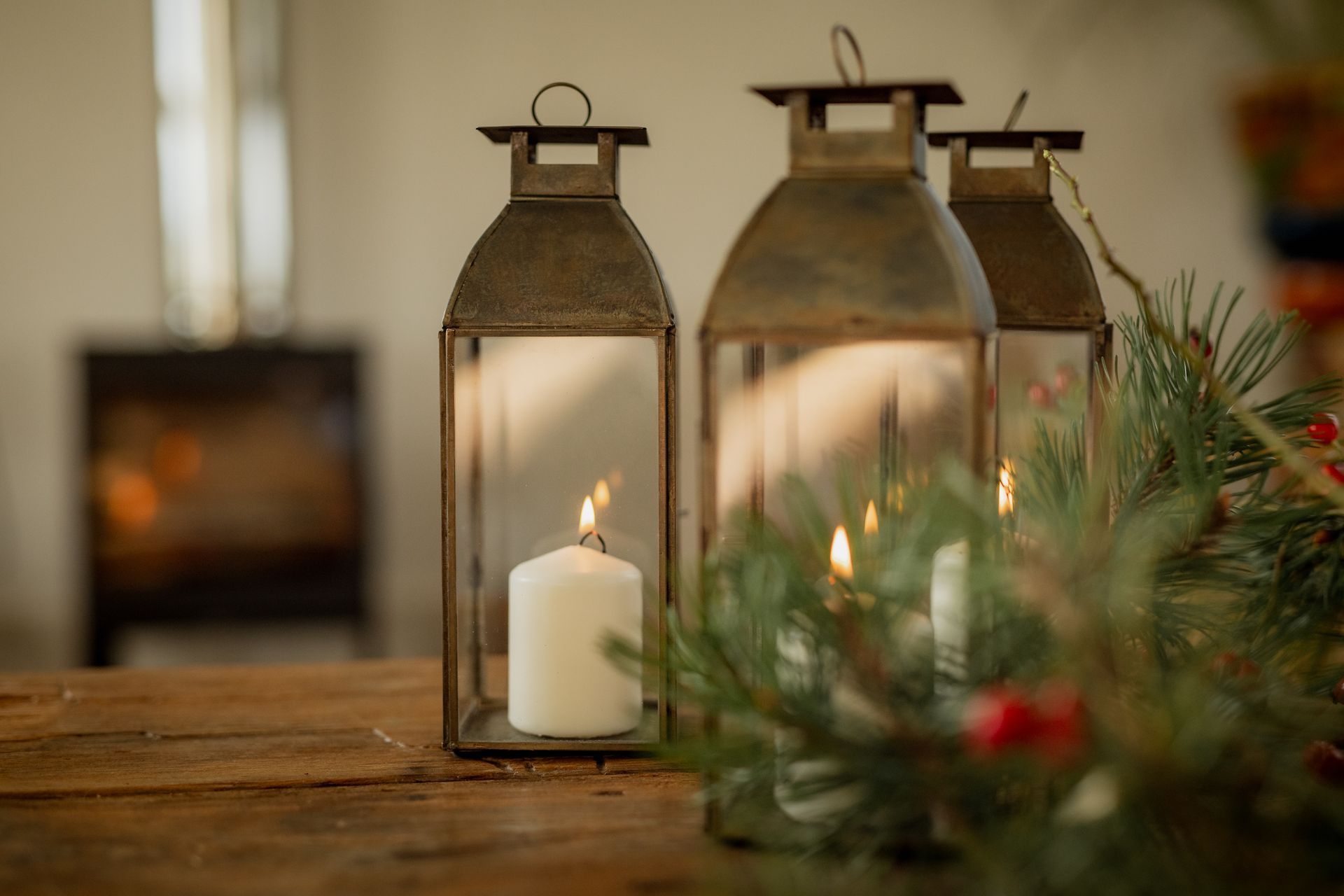 Three candle lanterns on a wooden table, with greenery and a fireplace in the background.