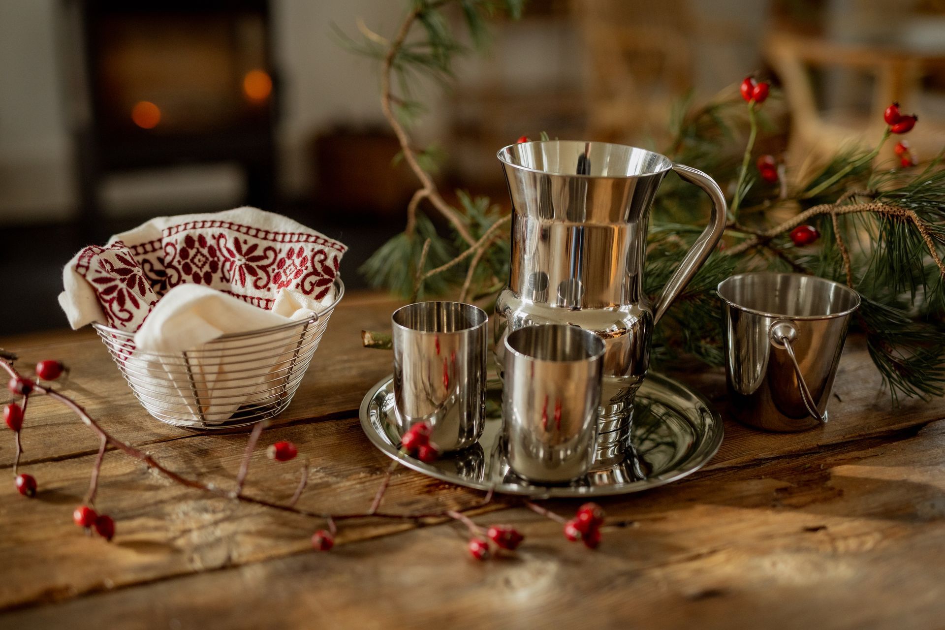 Silver serving set with pitcher, cups, and basket of bread on a wooden table, festive greenery.