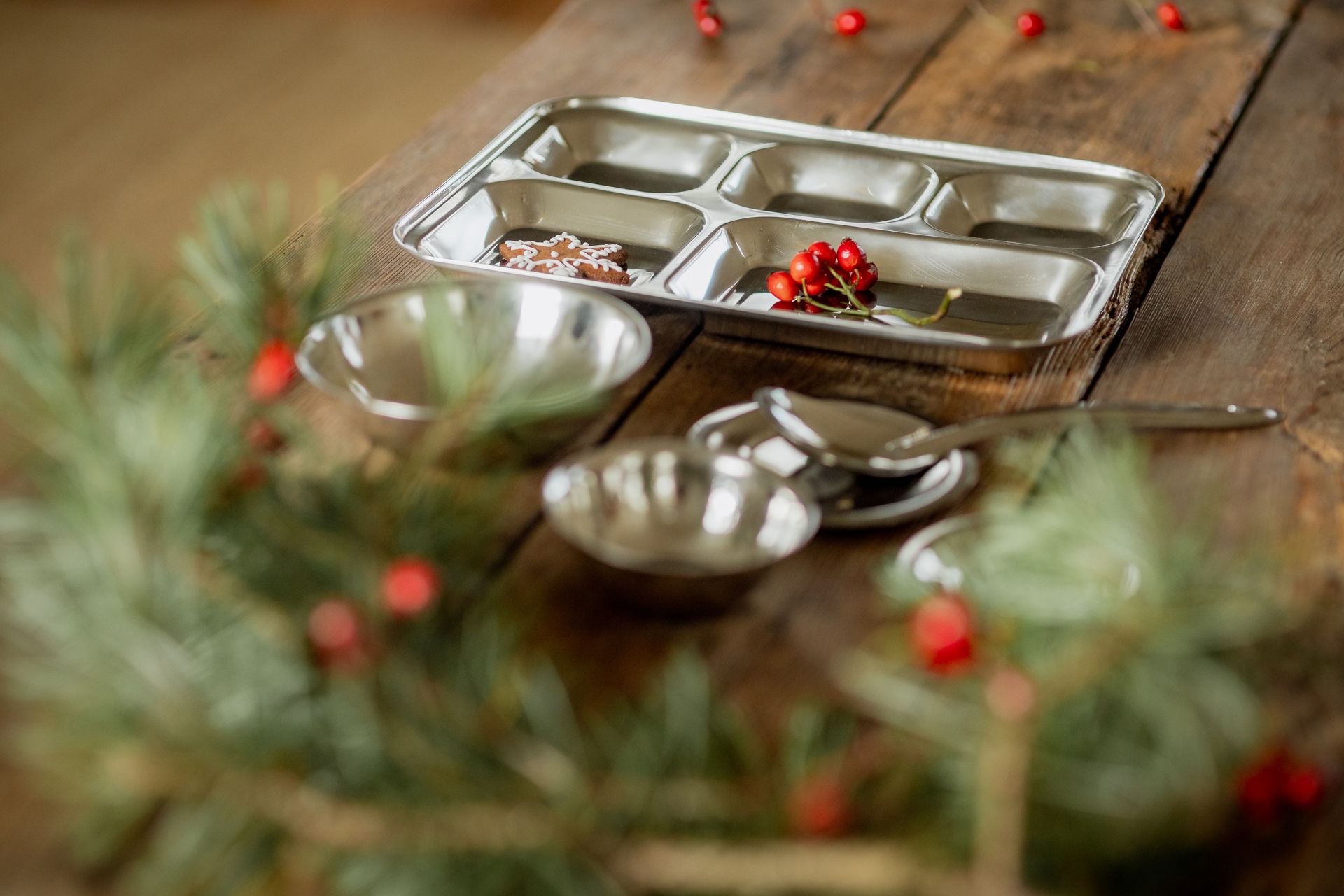 Stainless steel food tray with bowls and utensils on a wooden table, holiday greenery.