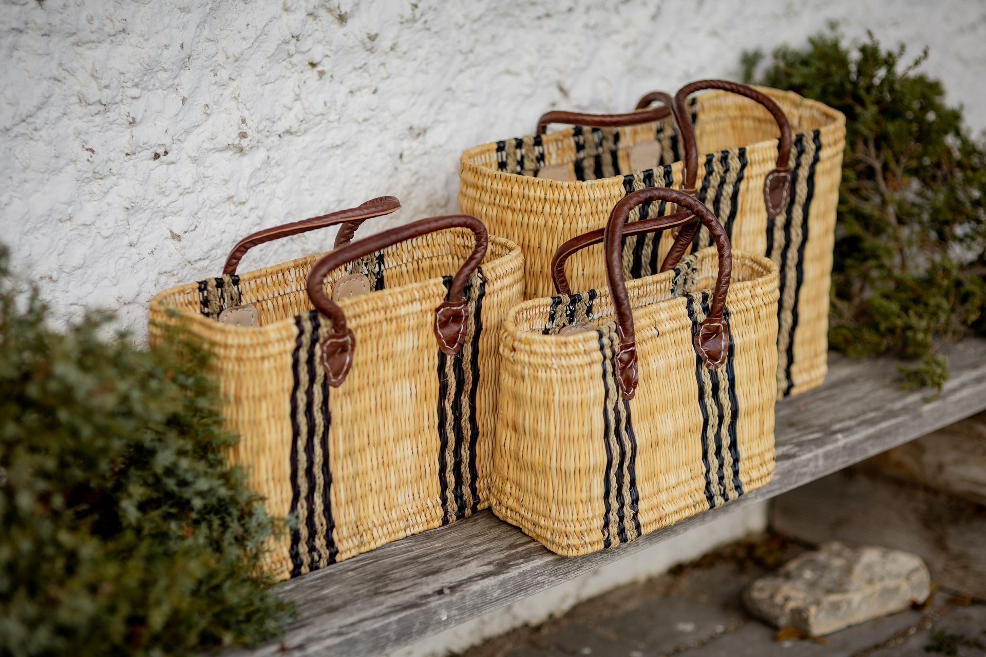 Four woven baskets with leather handles in varying sizes, lined up on a wooden bench, with greenery.