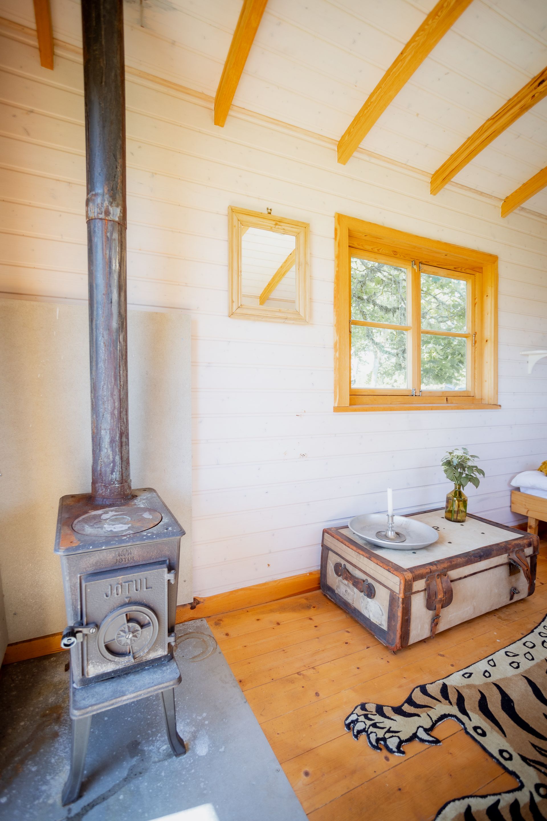 Cozy cabin interior with wood stove, trunk table, window, light-wood flooring, white walls, and tiger-print rug.