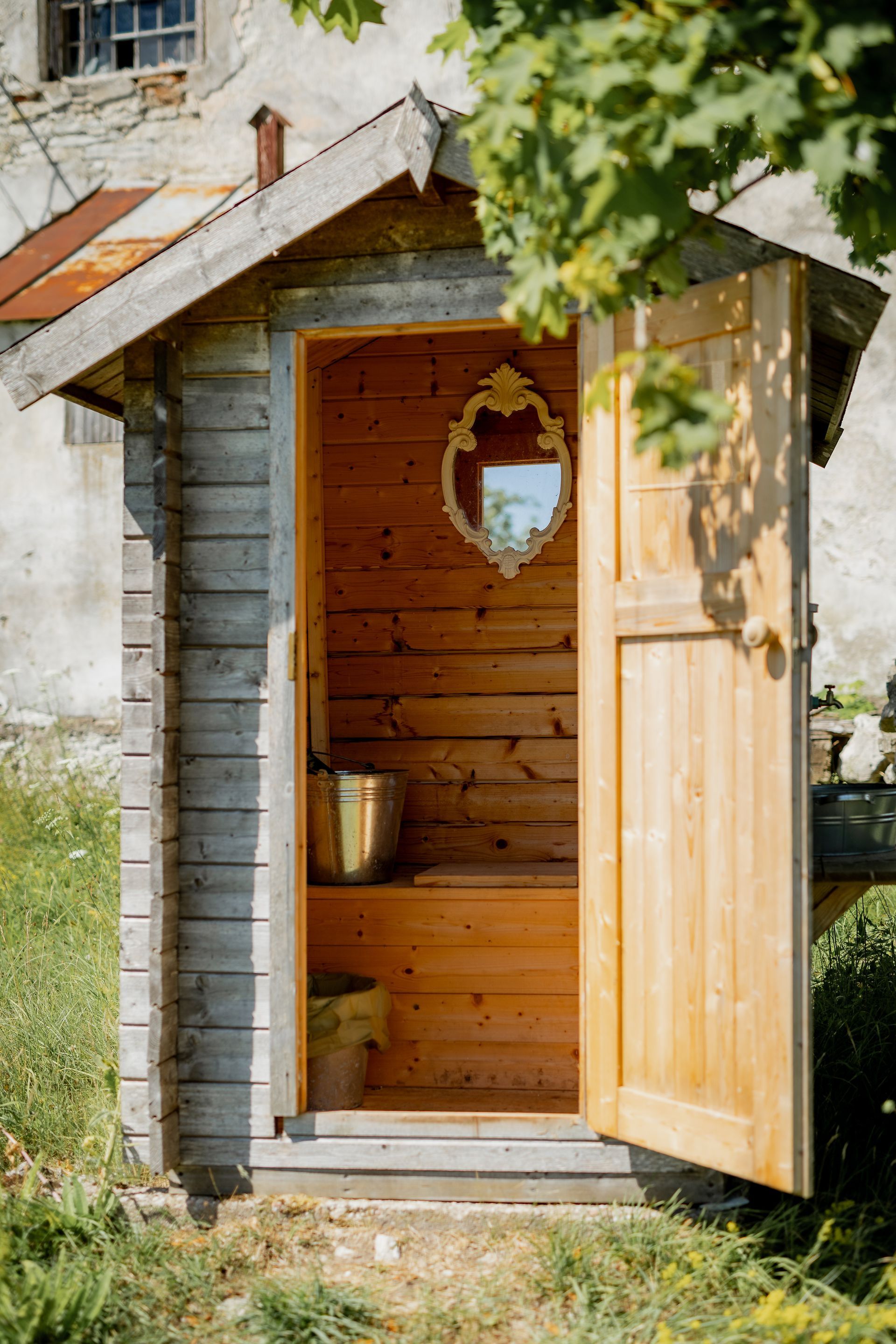 Rustic wooden outhouse with open door, gold mirror, and buckets inside. Building sits on grass near a stone building.