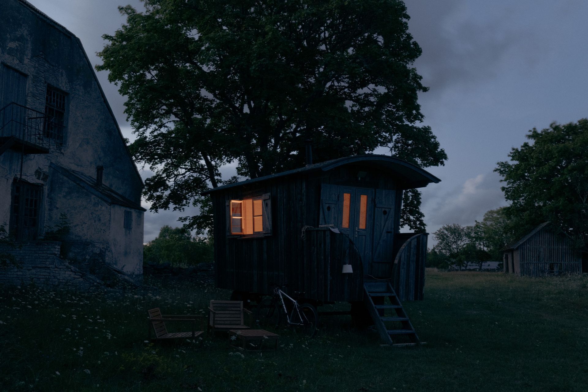Dark wooden shack with lit windows in a field at dusk. Wooden chairs sit in front.