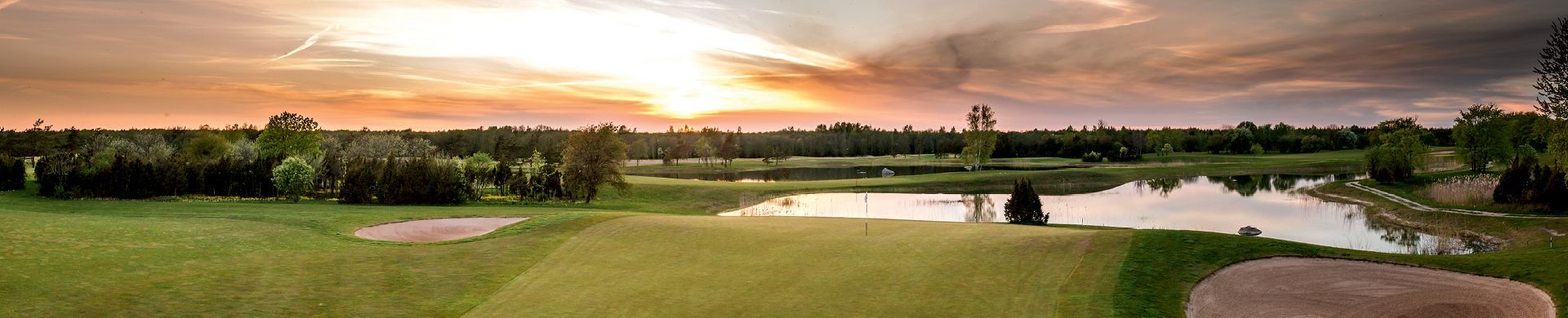 Golf course landscape with a lake at sunset. Green grass and trees frame the water and sky.