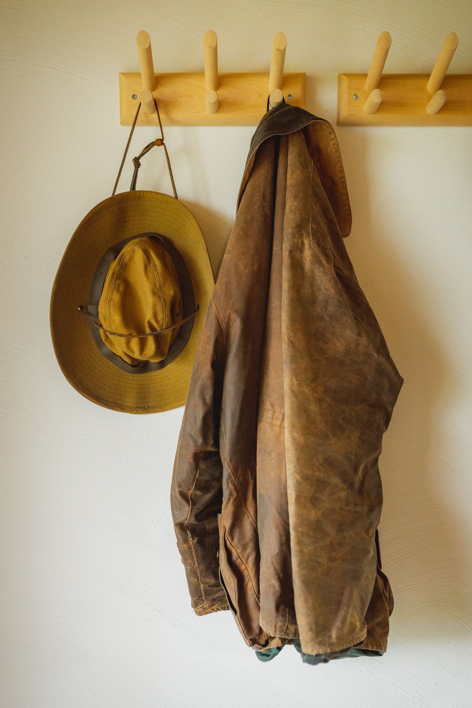 Tan hat and brown jacket hanging on wooden coat rack against white wall.