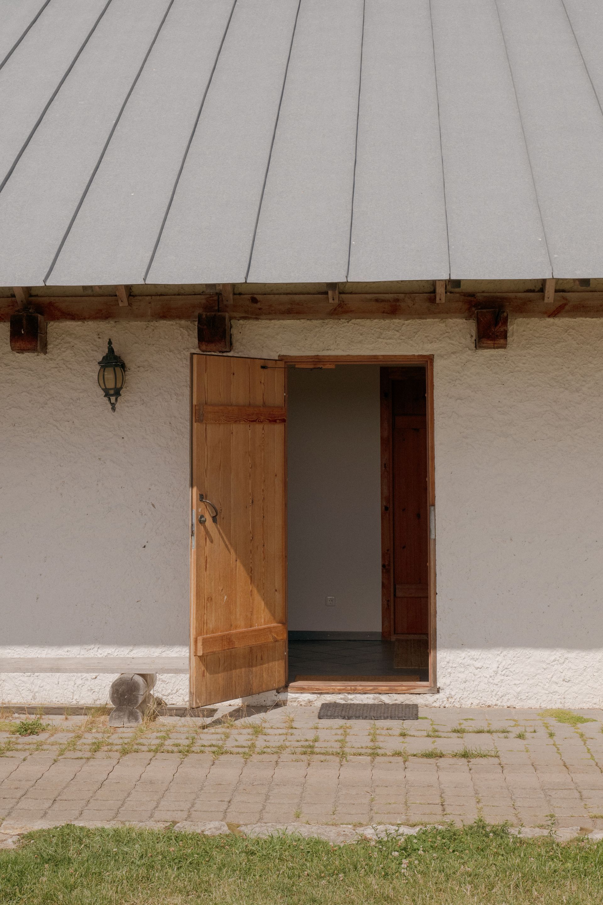 Wooden door of a white building open, revealing interior. Gray roof, brick walkway, and grass in the foreground.