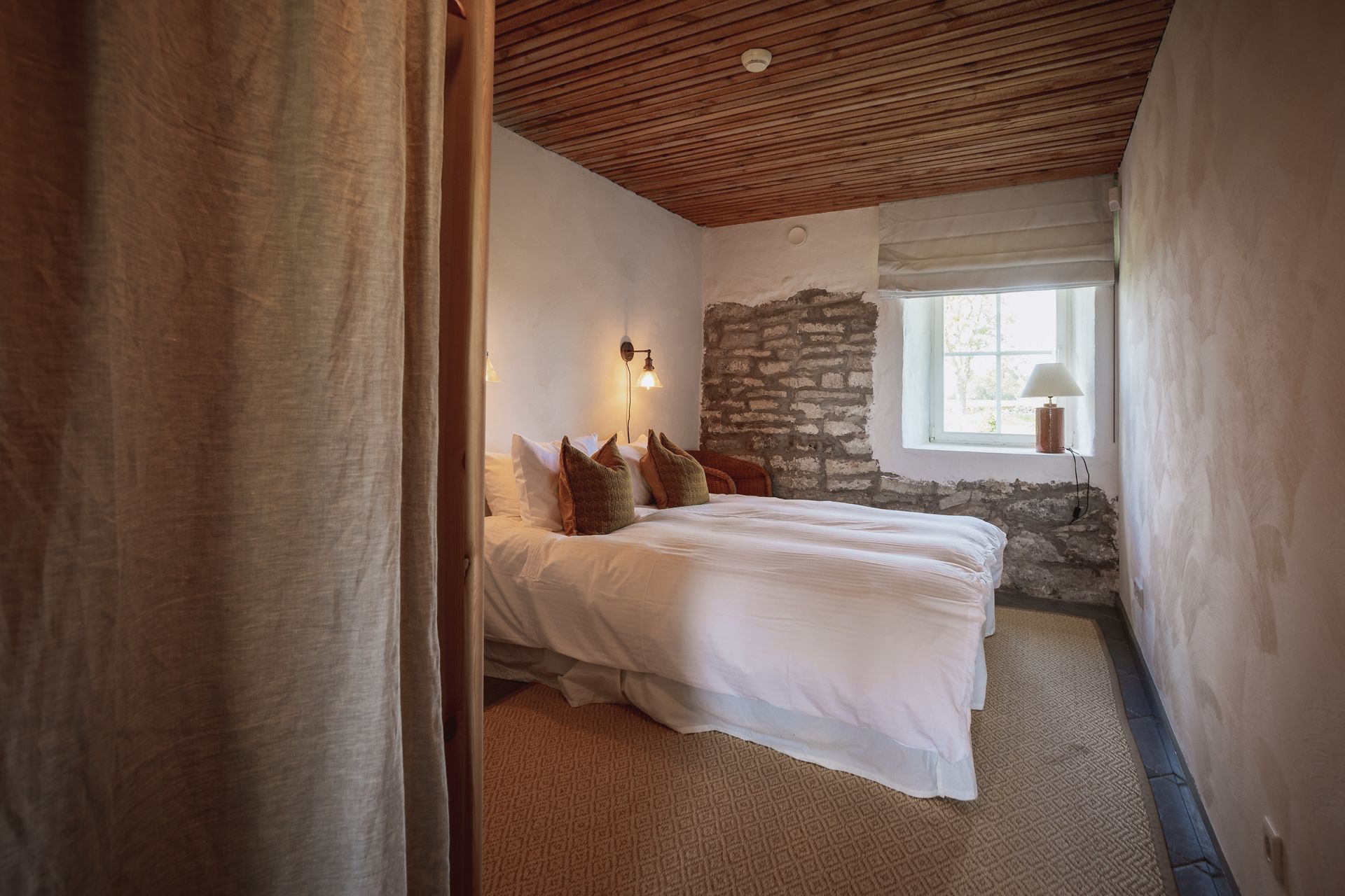 Bedroom with a bed, exposed stone wall, wood ceiling, and a window with a lamp.
