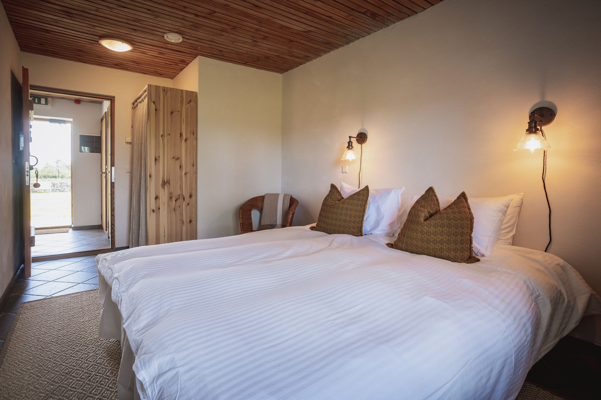 Bedroom with a double bed, wooden ceiling, and side lamps. An open door leads to another room.
