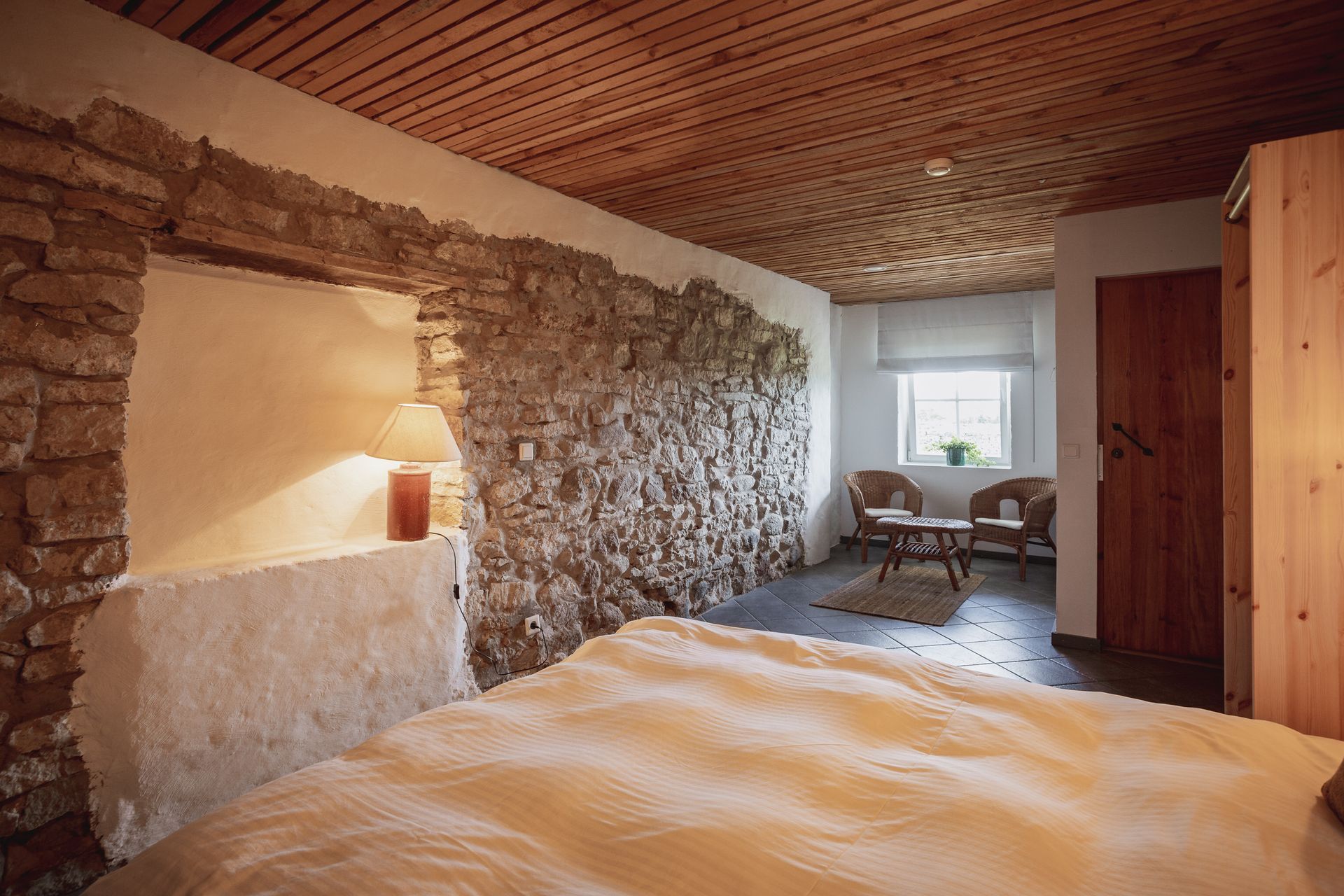 Bedroom with stone wall and wooden ceiling, bed in foreground, small seating area by window.