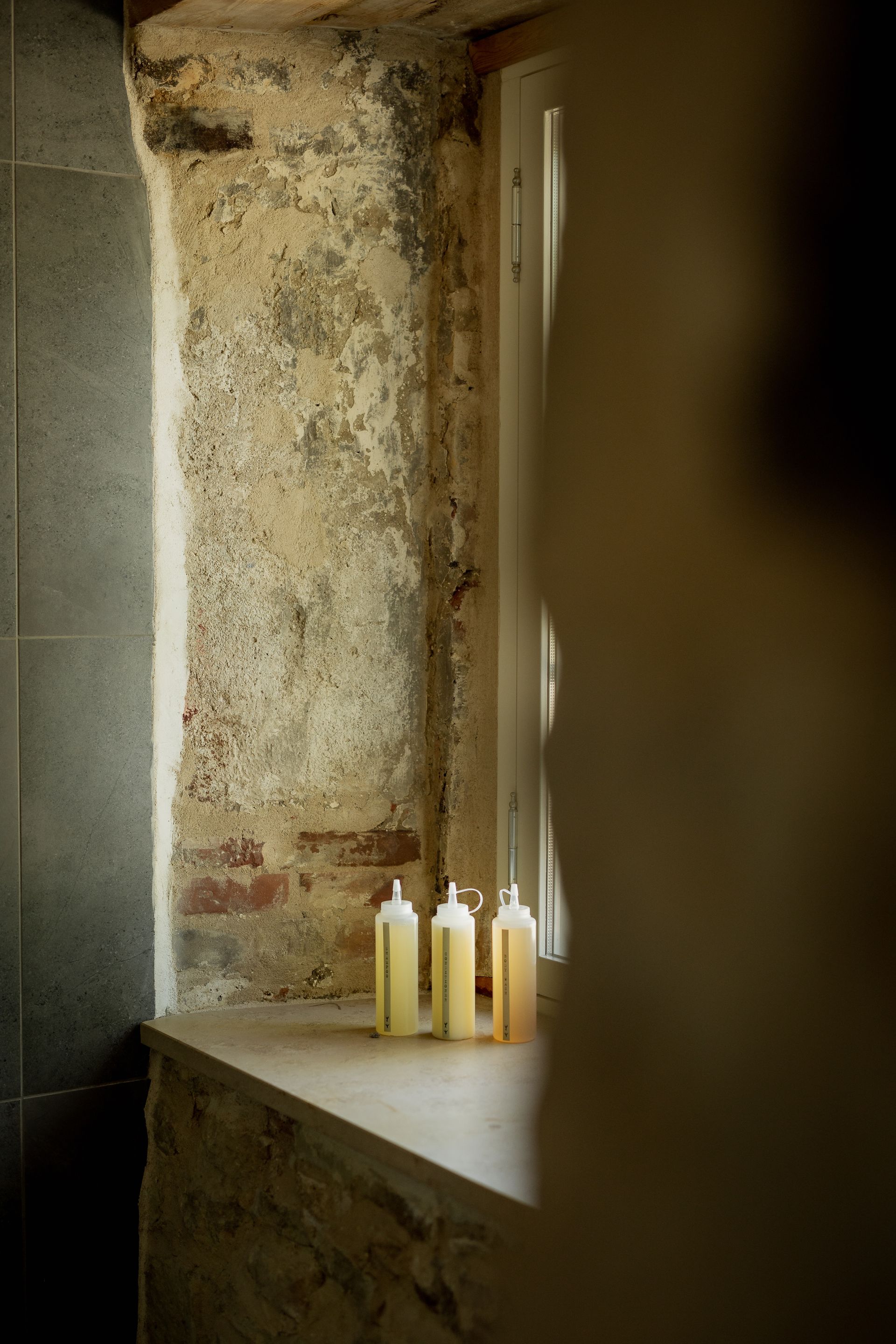 Bottles of body wash on a stone windowsill next to a window. The wall is aged and worn.