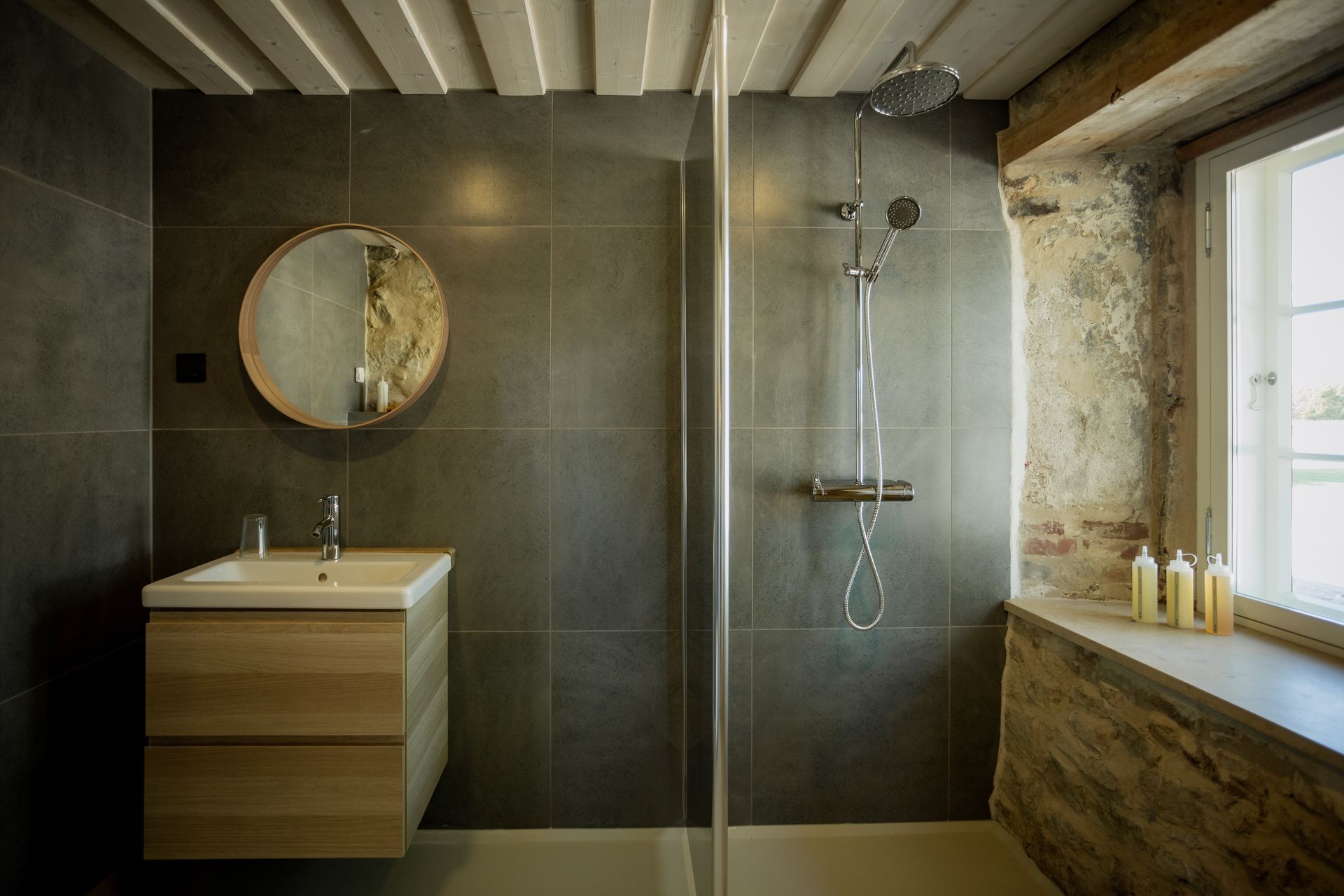 Bathroom with grey tiled walls, glass shower, wooden vanity, round mirror, and stone window frame.