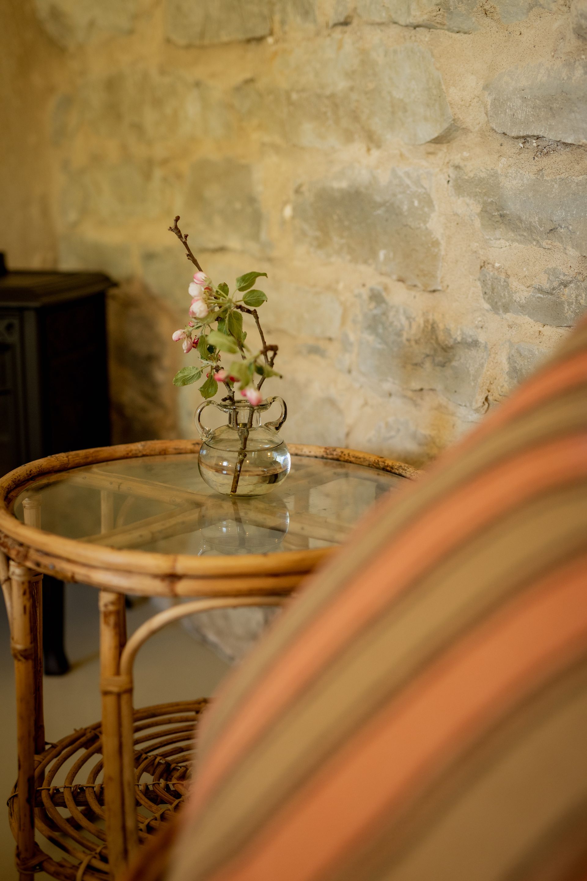 A small floral arrangement on a round, wicker table in front of a stone wall, with part of a striped cushion visible.