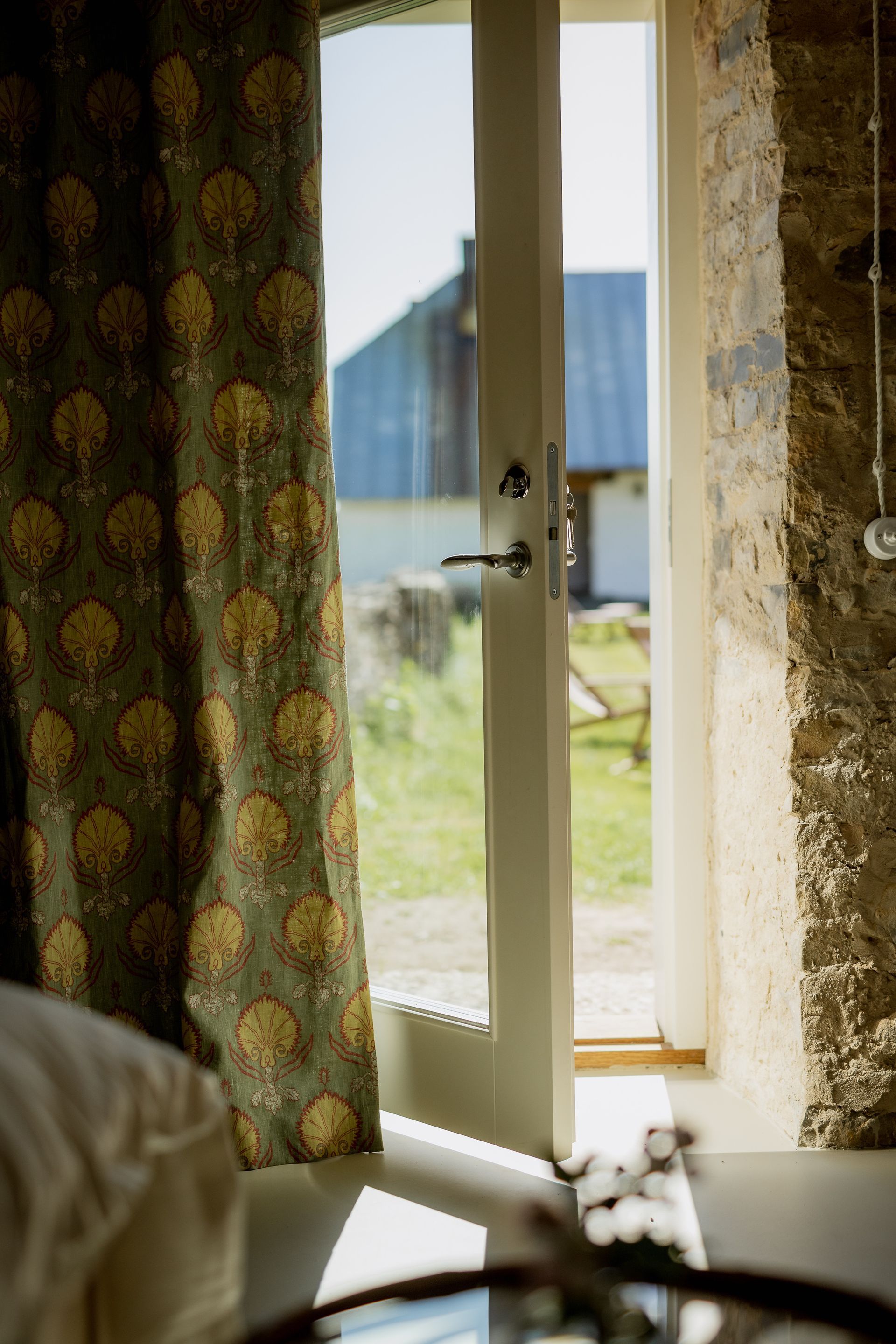 Open white door reveals a grassy yard and building in sunlight, framed by a patterned curtain and stone wall.