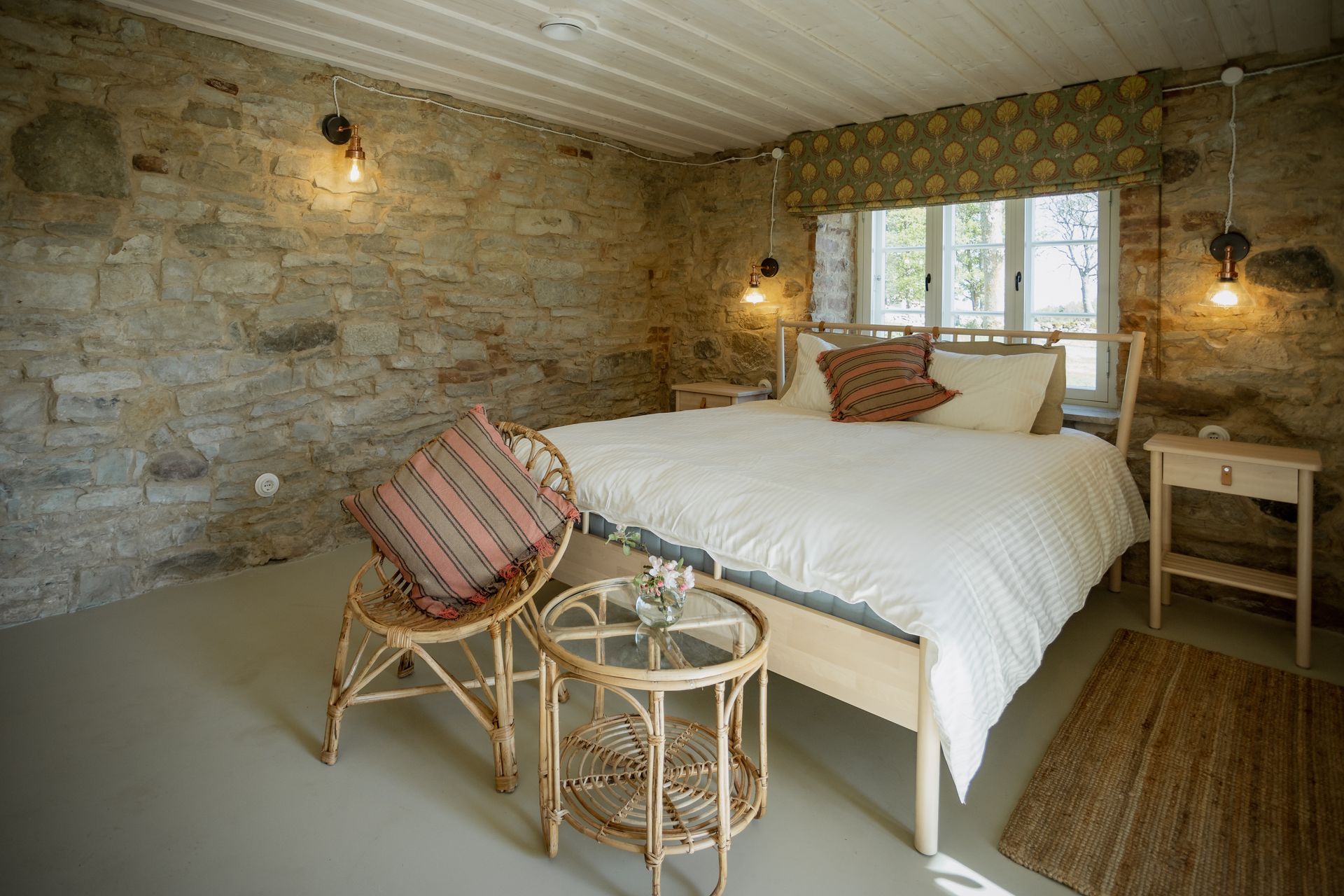 Bedroom with stone walls, light wood bed frame, white bedding, woven chair, and small tables.