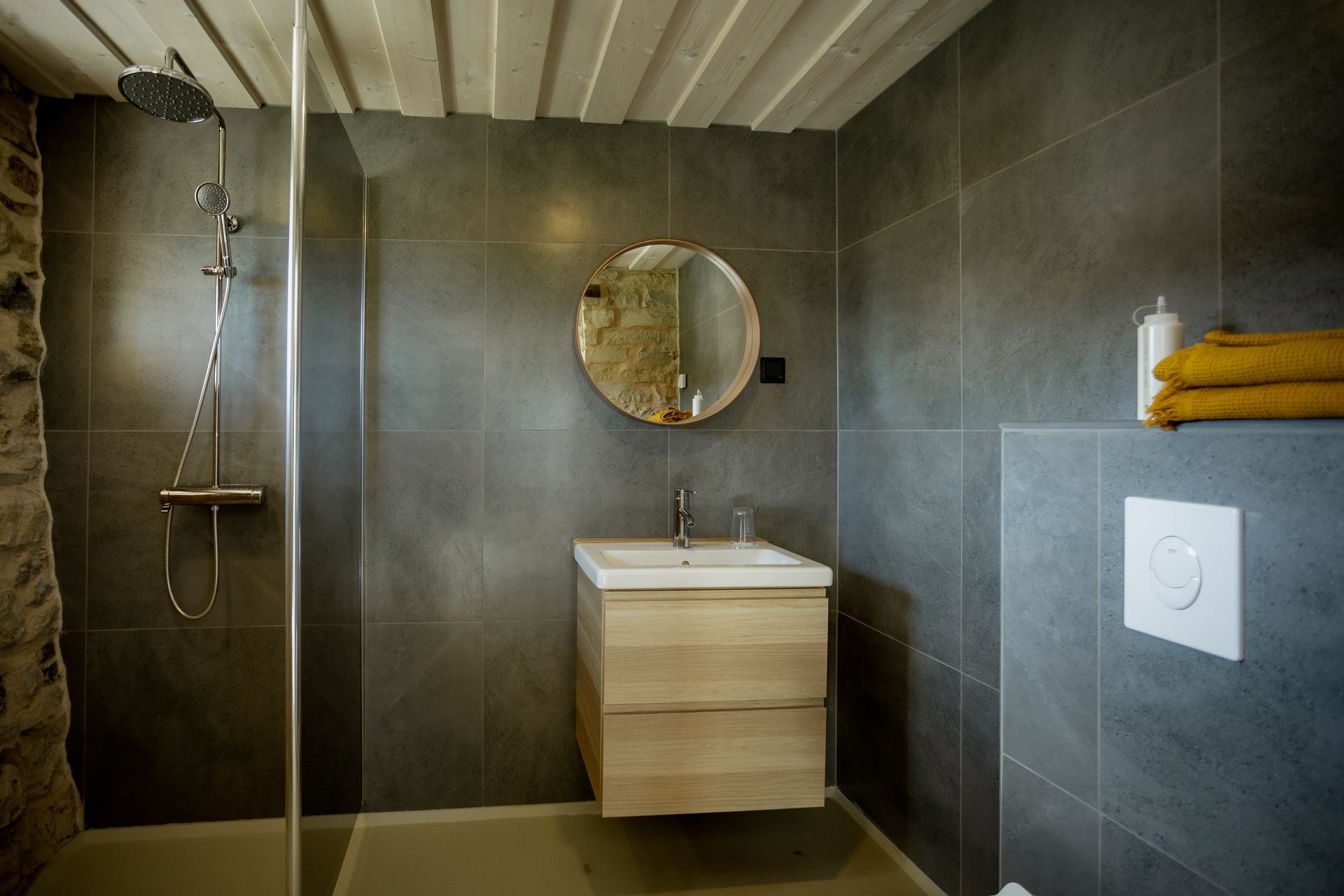 Bathroom with stone wall and gray tile, shower, wooden vanity, round mirror, and towels.