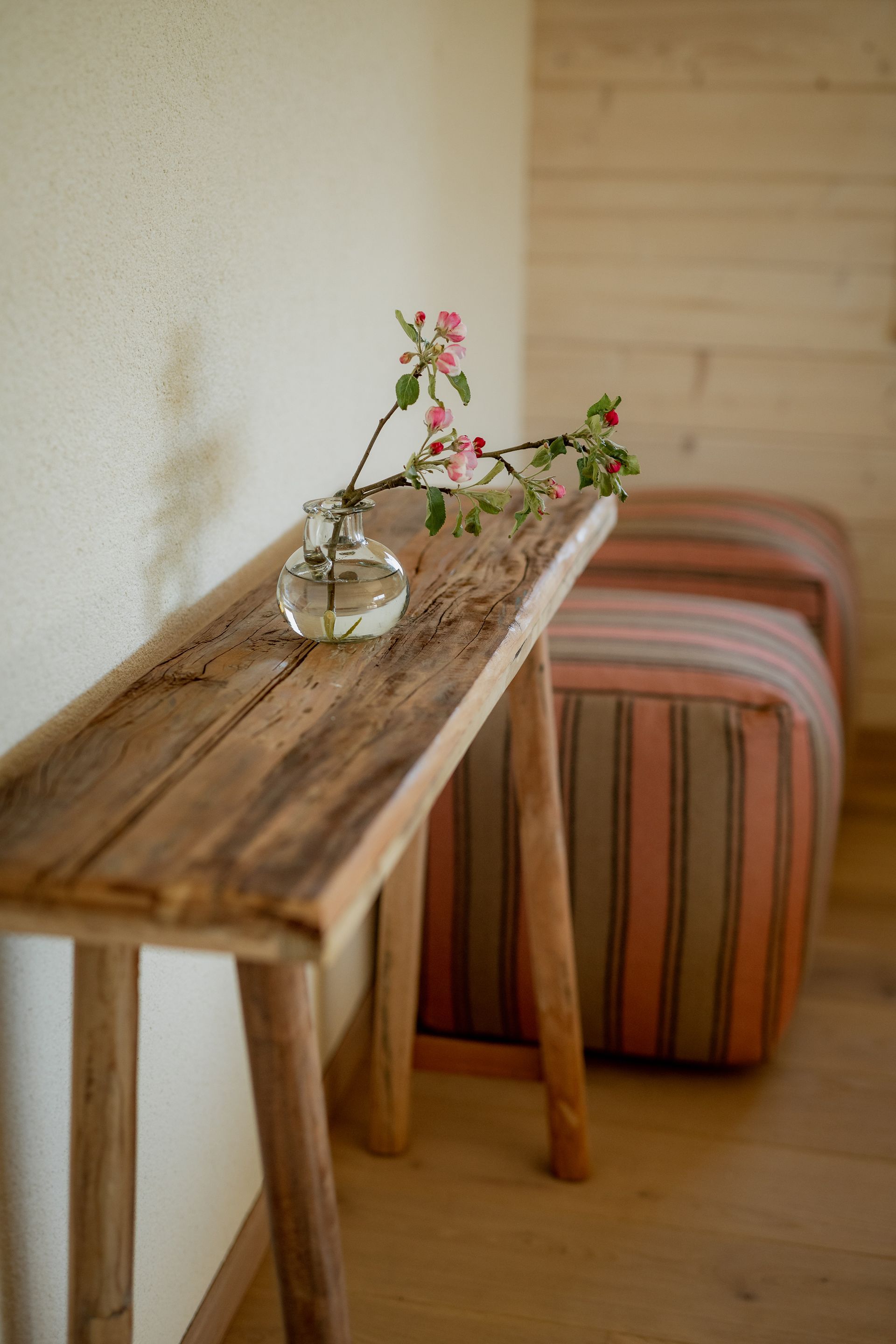 Wooden bench with a vase of pink flowers; next to a striped ottoman.