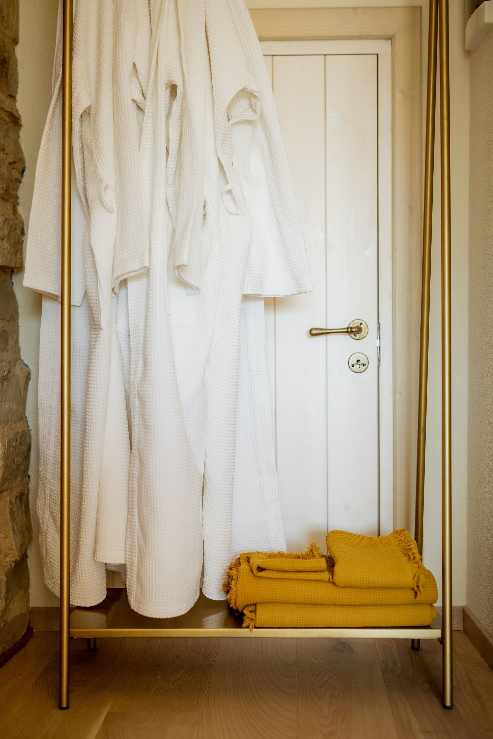 White bathrobes hanging on a gold rack, with folded yellow towels below. A white door is in the background.