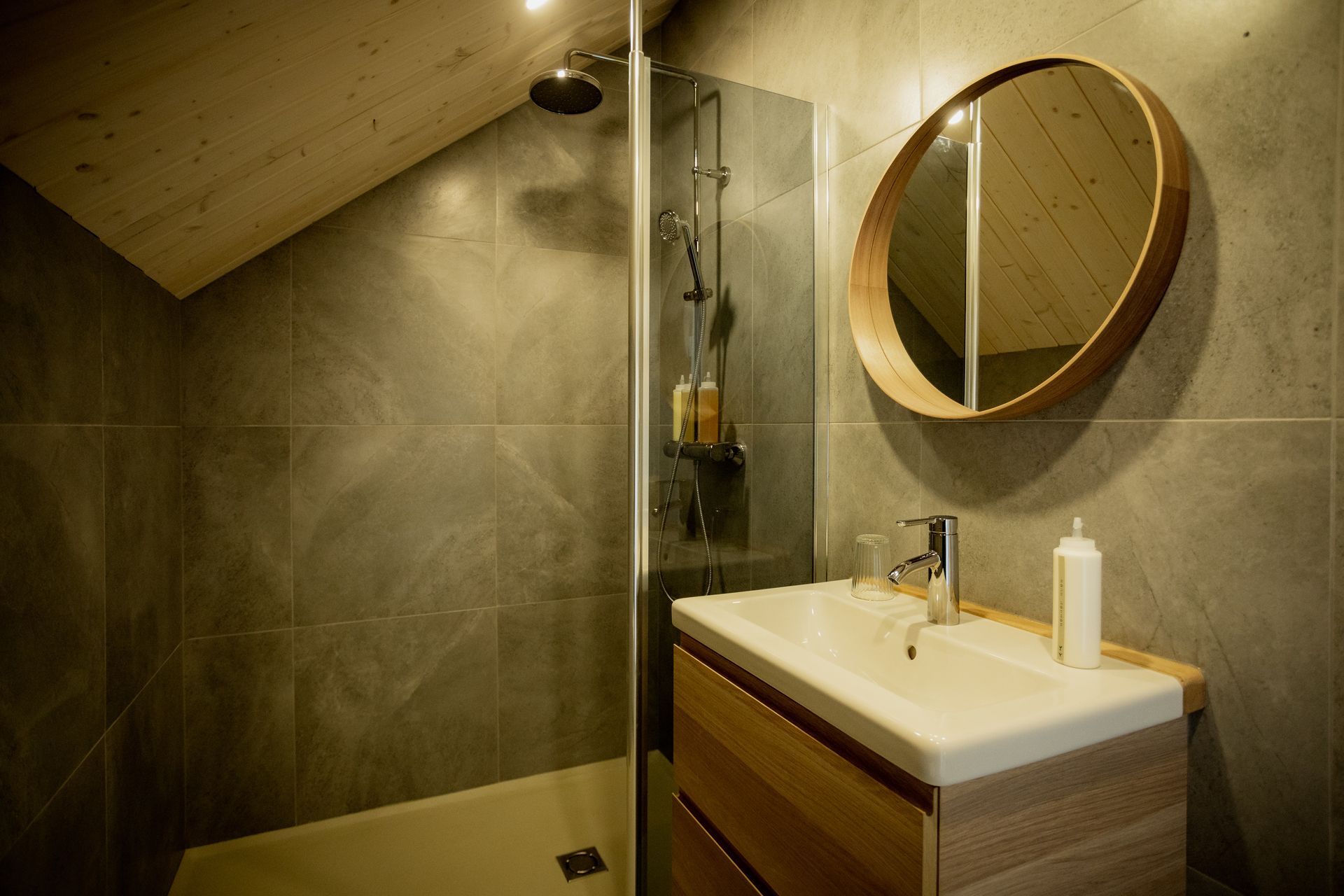 Bathroom with gray tile walls, wooden vanity, round mirror, and a glass shower.