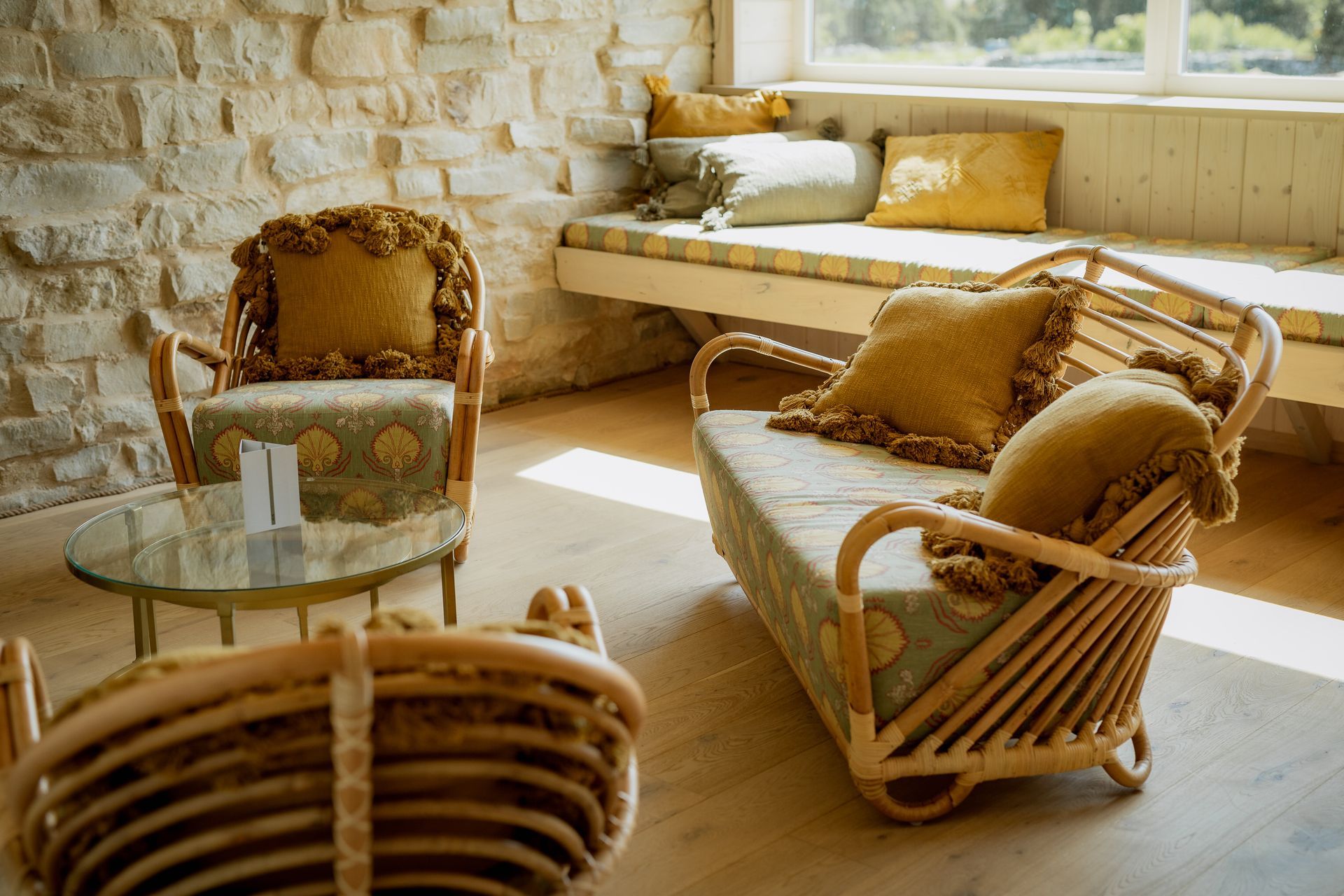 Rattan furniture in a sunlit room: sofa, chairs, round table, stone wall, window seat with pillows.