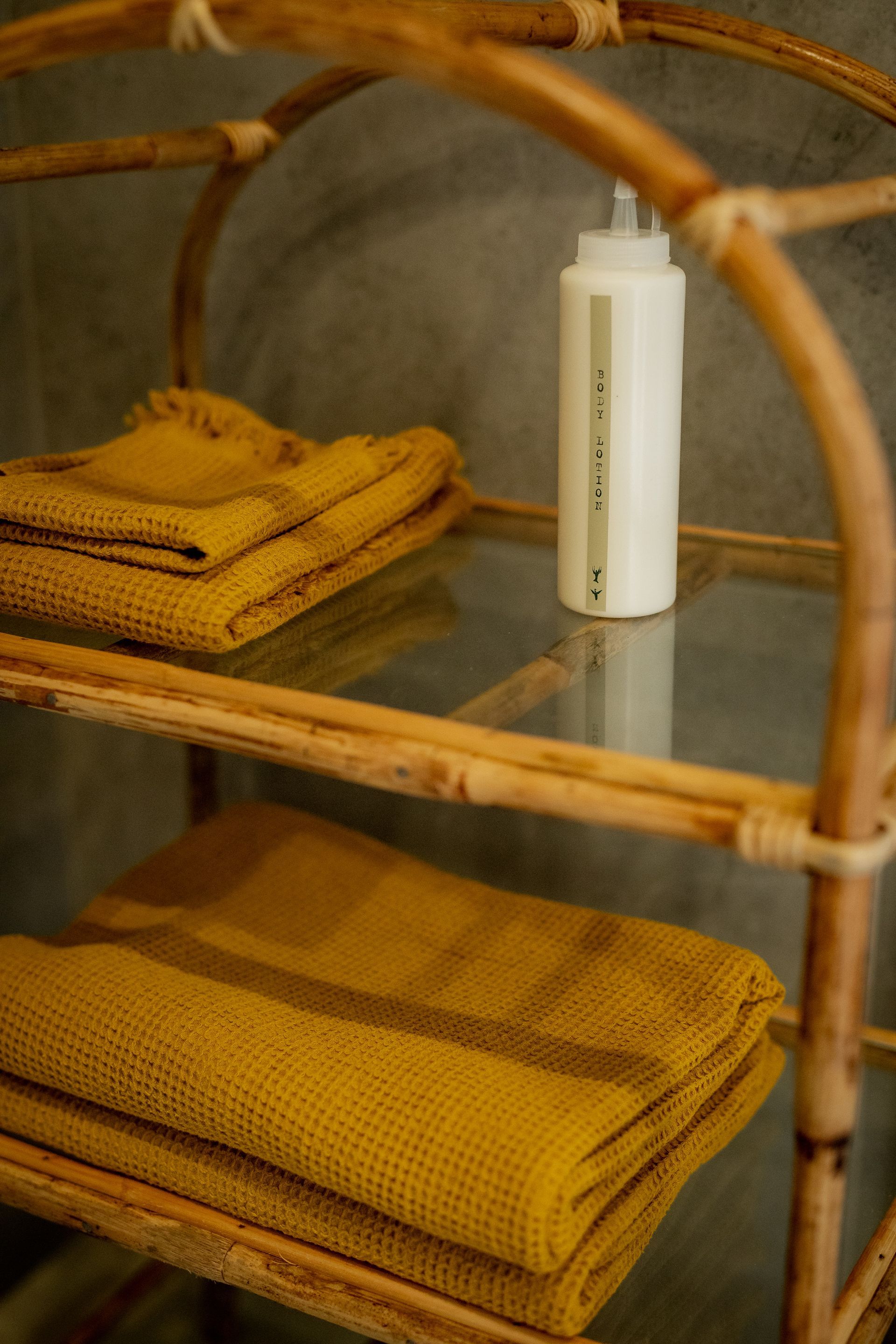 Rattan shelving unit with folded golden towels and a white lotion bottle on a glass shelf.