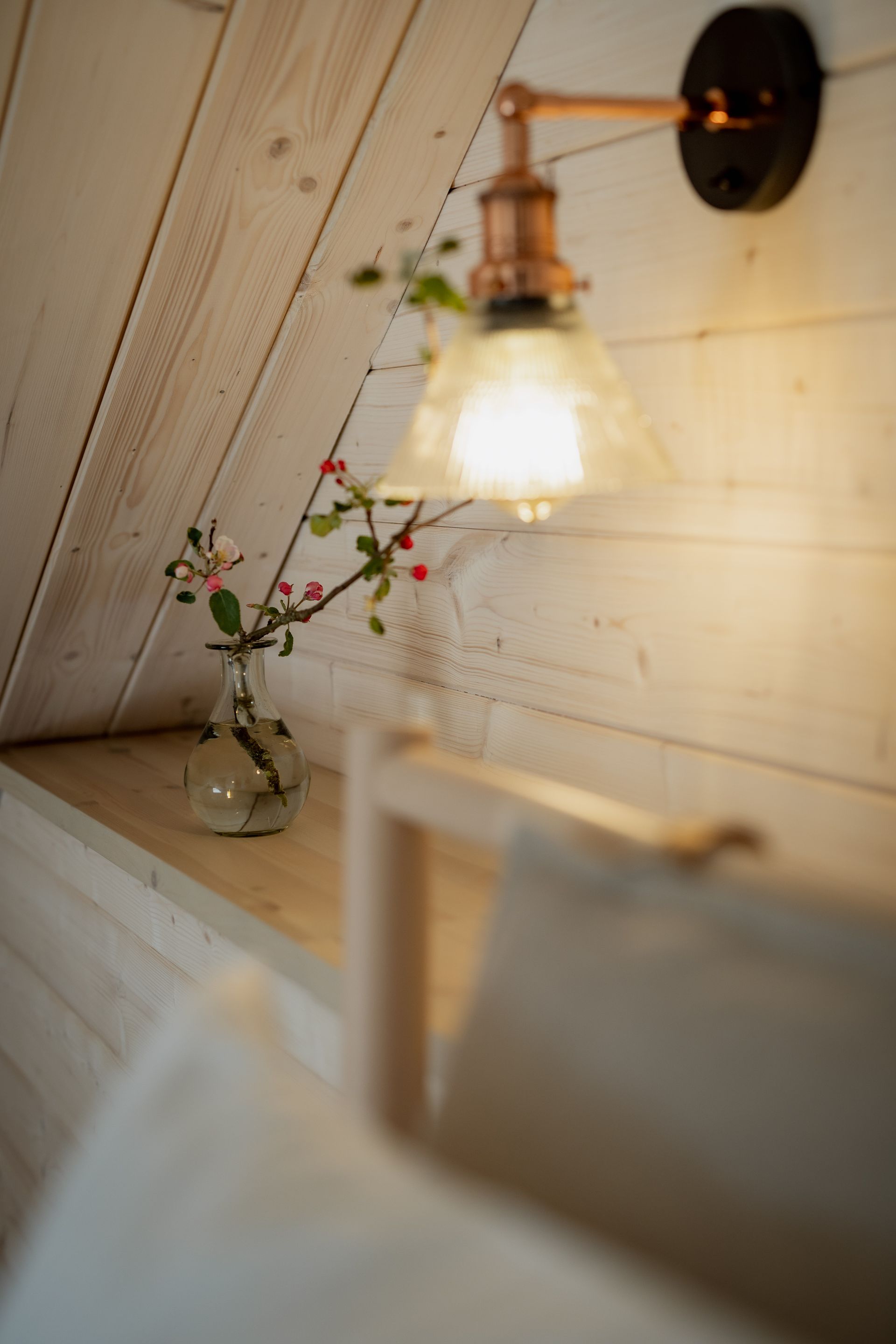 Cozy bedroom with wooden walls, lit by a copper-toned wall lamp. A vase with red berries sits on a shelf.