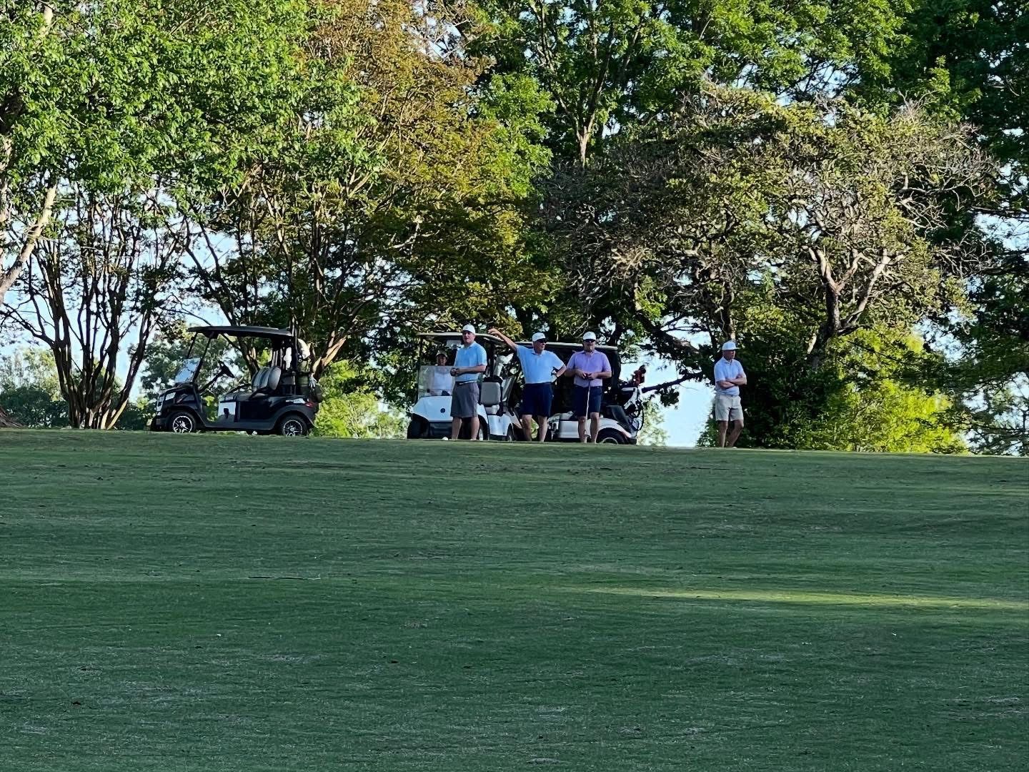 Golfers standing on a green, near golf carts, under trees. It's a sunny day.