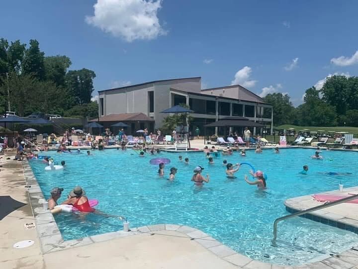 A crowded outdoor swimming pool on a sunny day. People are swimming, playing, and sunbathing near a two-story building.
