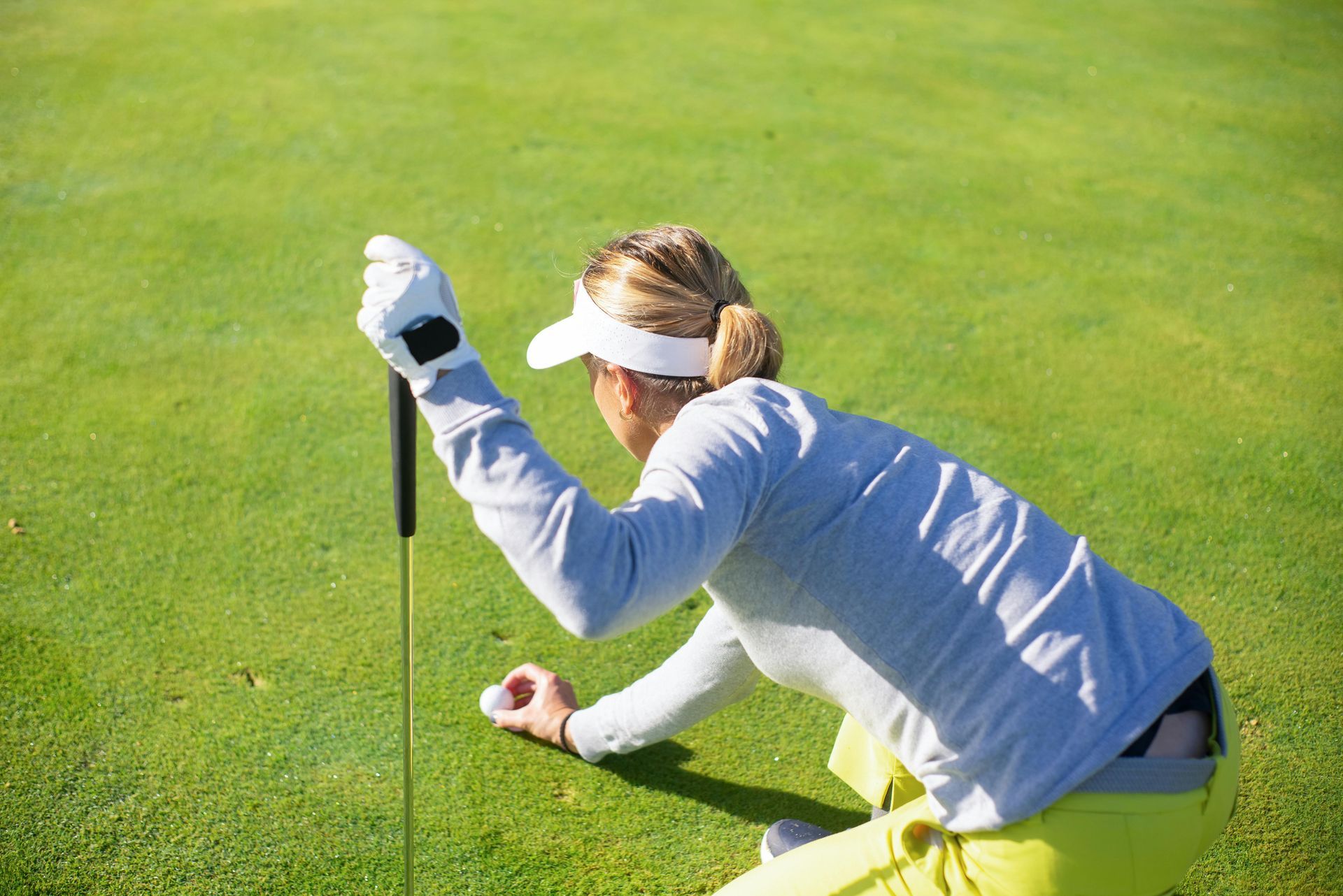 Woman on a golf course lining up a putt, wearing a visor, grey top, and yellow shorts.