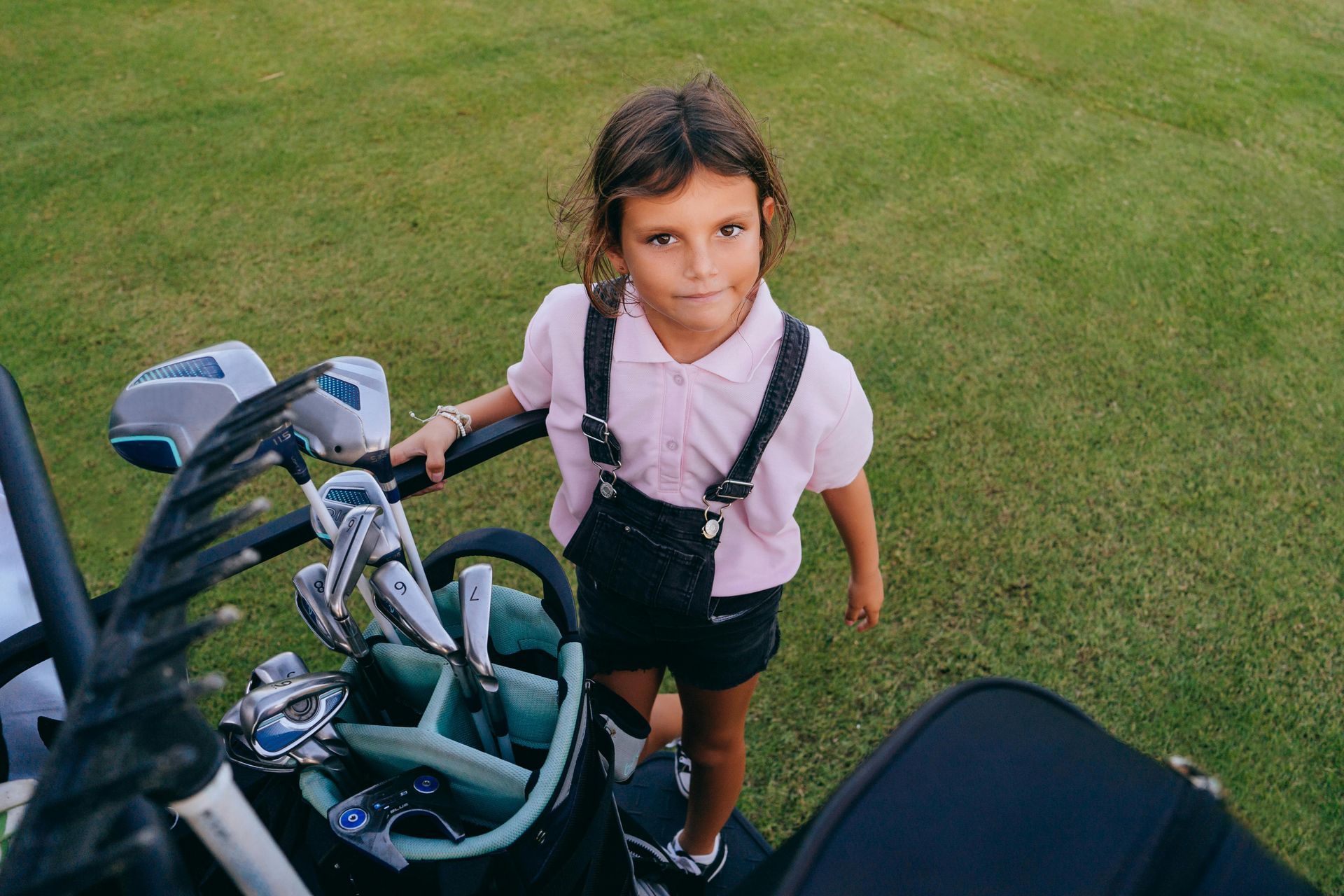 Young girl standing next to golf bag on golf course, wearing pink shirt and overalls.