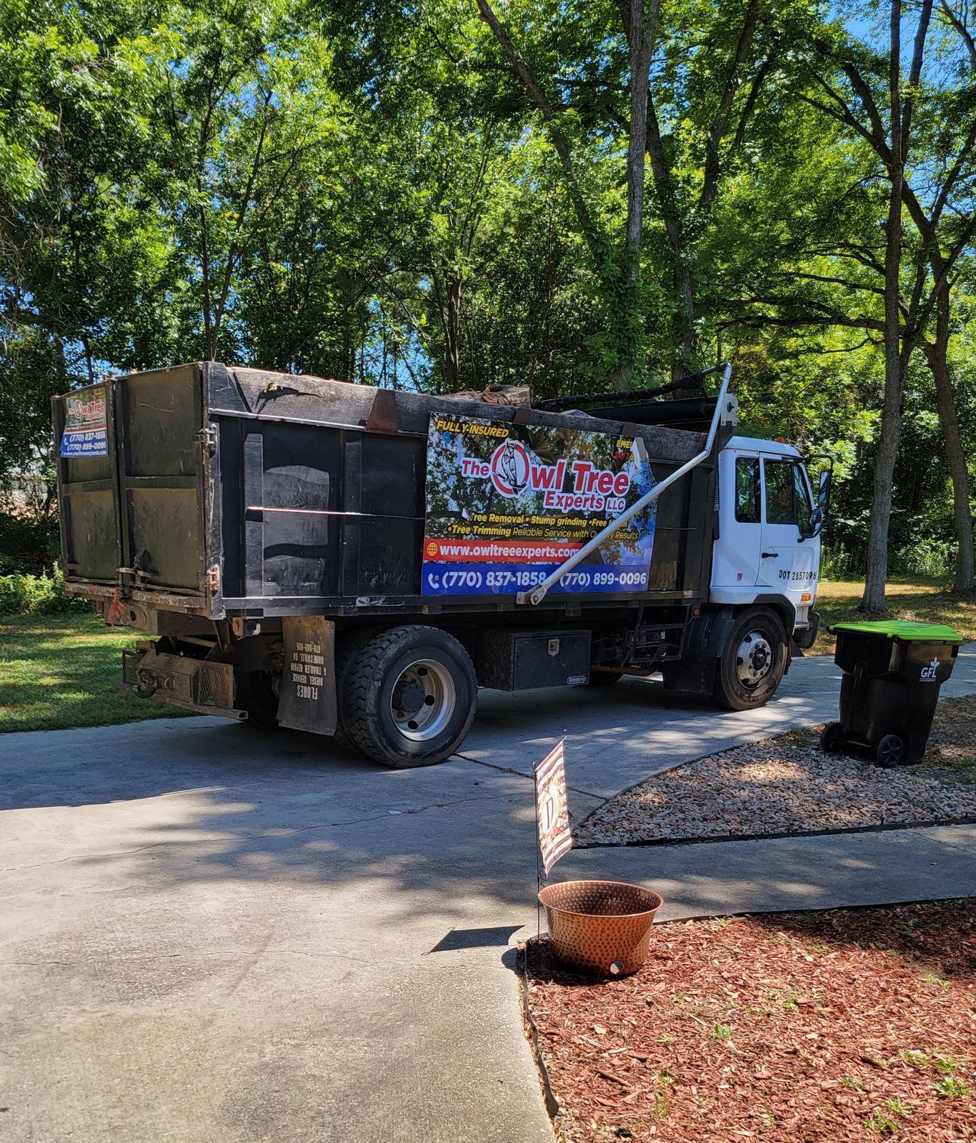 Dump truck parked on a driveway, full of debris. A trash can sits nearby. Sunlight shines.