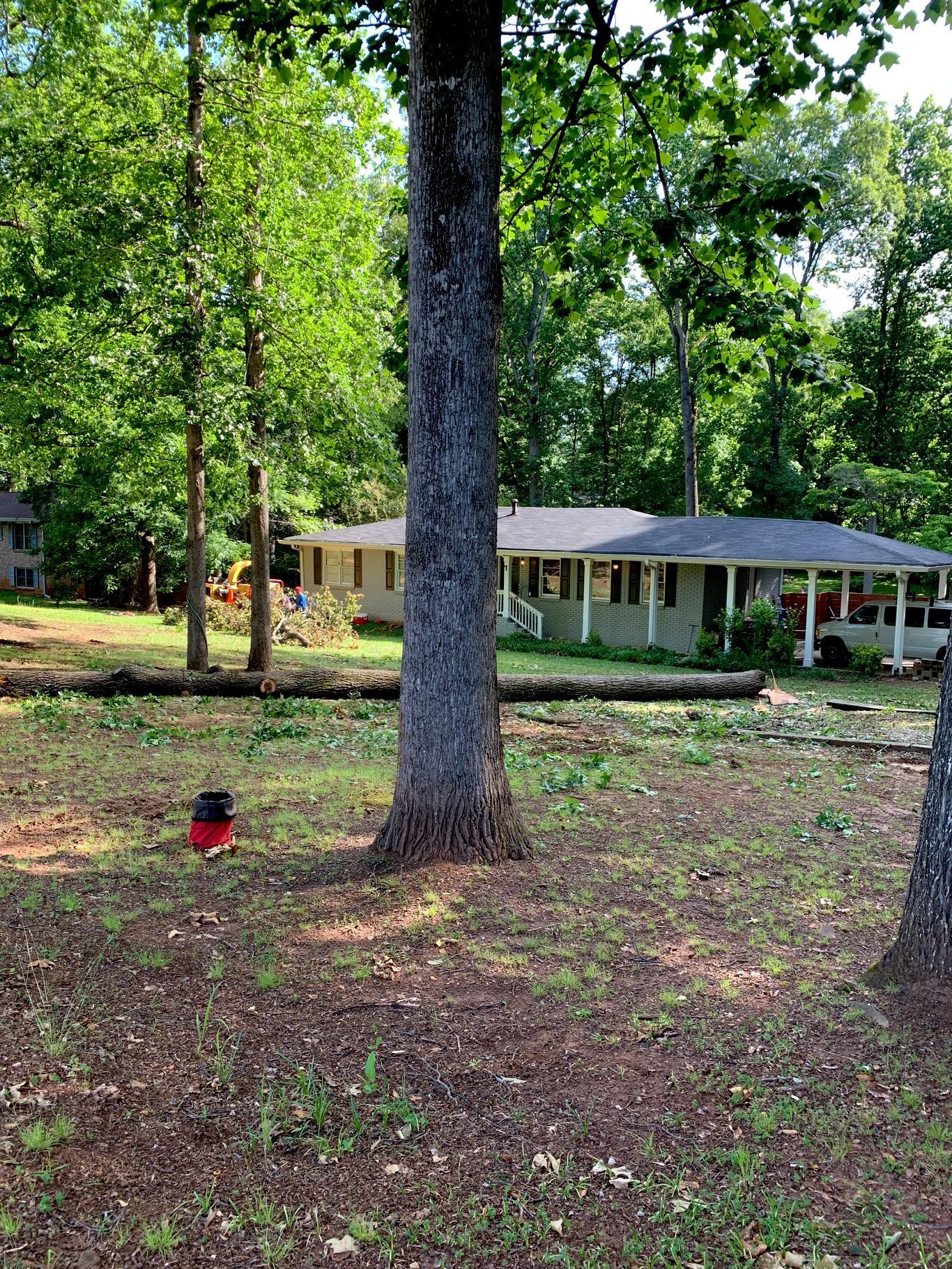A house with a porch is surrounded by trees in a wooded area. A red bin sits in the yard.