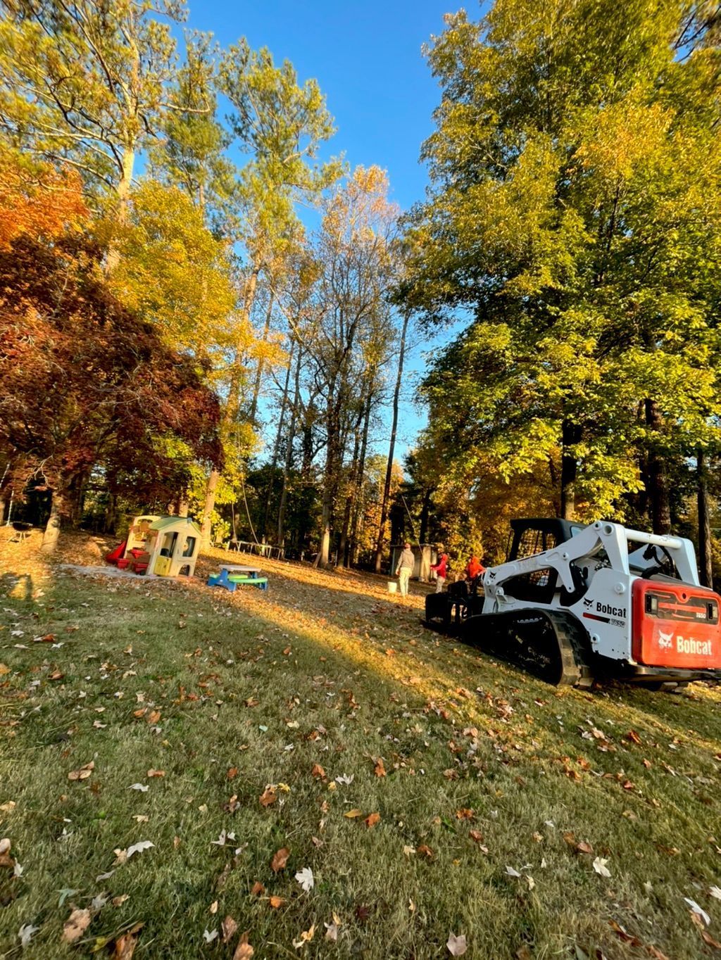 A Bobcat skid steer on a grassy area, trees with fall foliage in background.