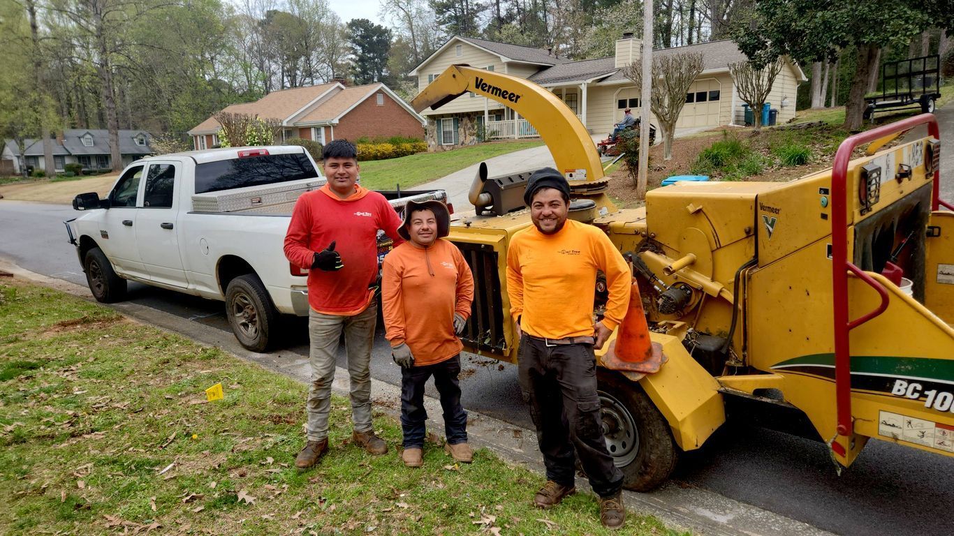 Three people stand next to a wood chipper and truck on a residential street.