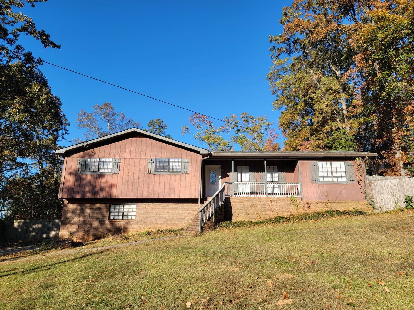 Brown split-level house on a grassy hill, surrounded by trees with autumn foliage, under a blue sky.
