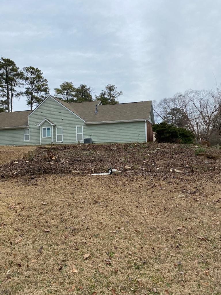 A house with pale green siding sits on a slightly elevated, mulch-covered landscape. Brown grass is in the foreground.