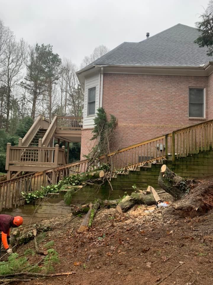 Person cutting logs on a hillside near a house with a wooden deck and stairs.