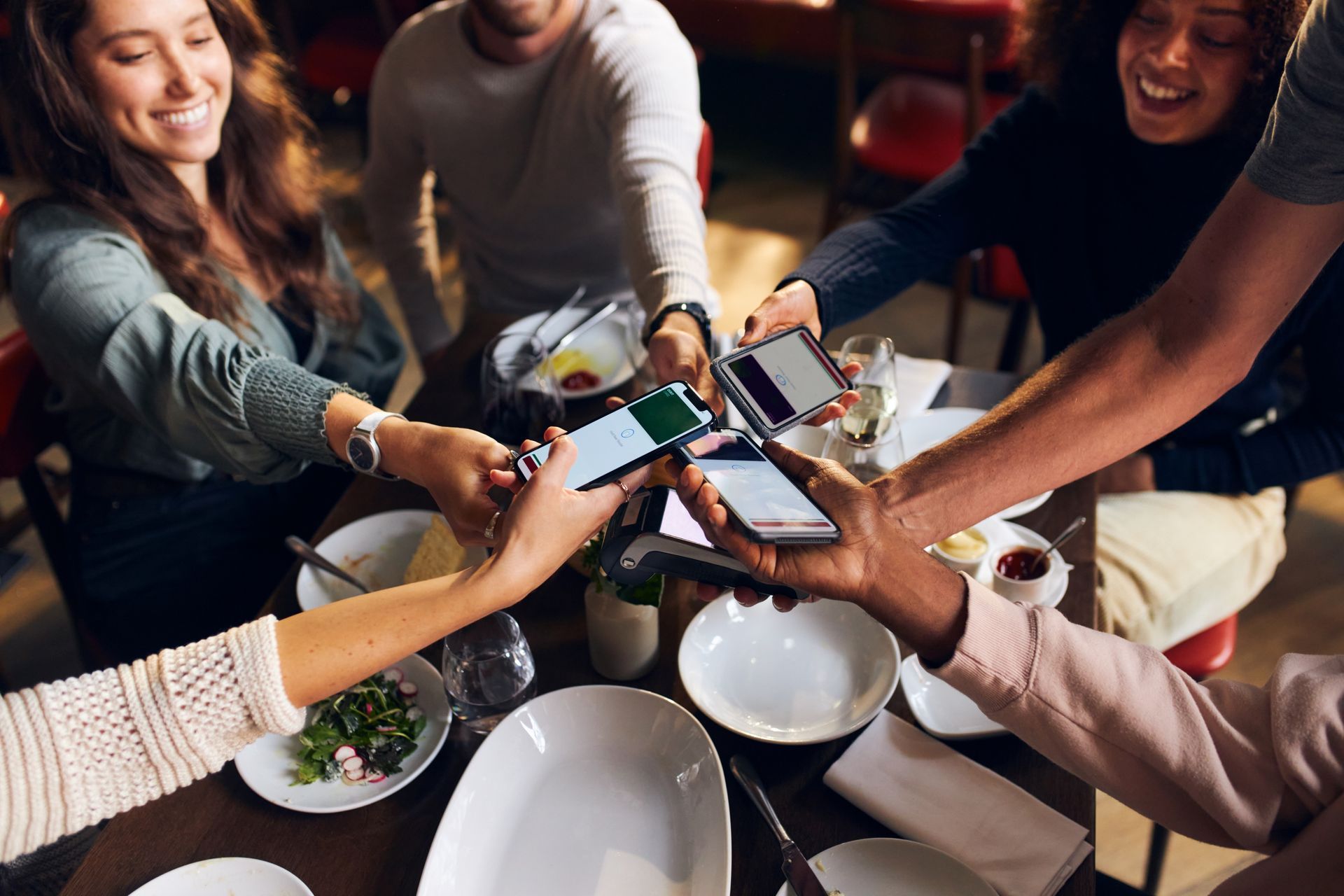 Group of people at a restaurant, holding phones up. Smiling and interacting over the table of food.