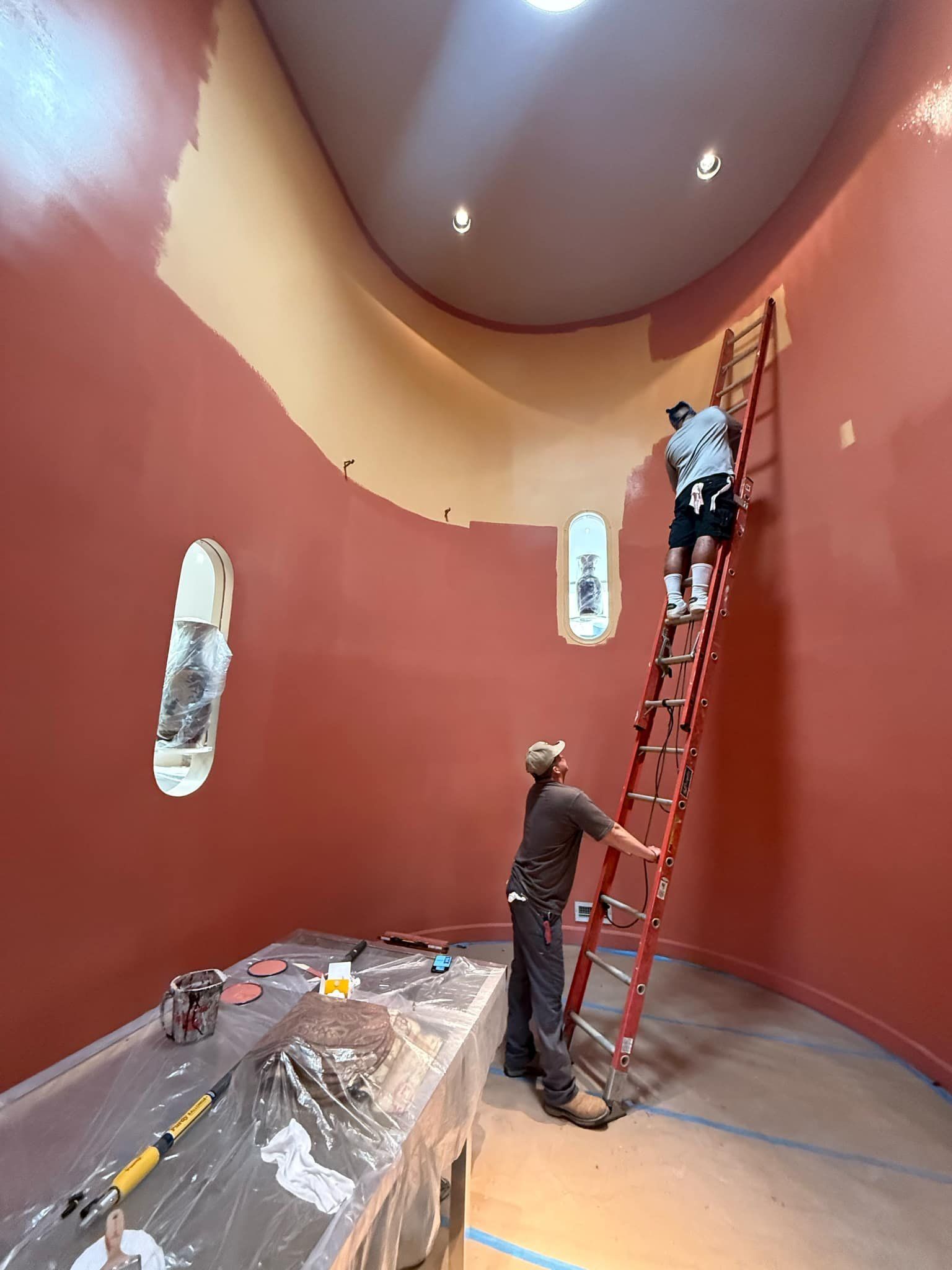 Two people painting a curved room. One on a ladder, the other at its base. Red and beige walls.